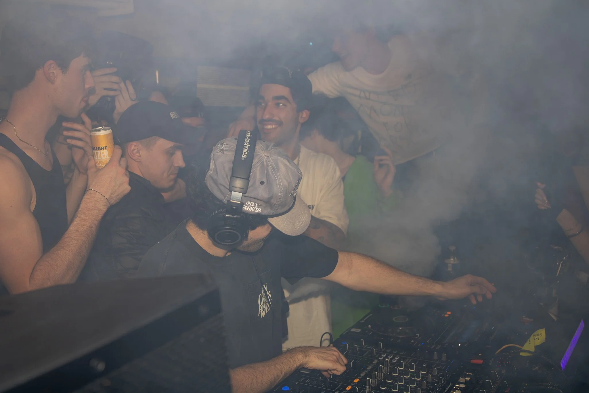 A DJ wearing a baseball cap and headphones performs on a mixing console at a party or nightclub, surrounded by smiling and engaging young people with some holding drinks and taking photos, amidst smoke or fog.