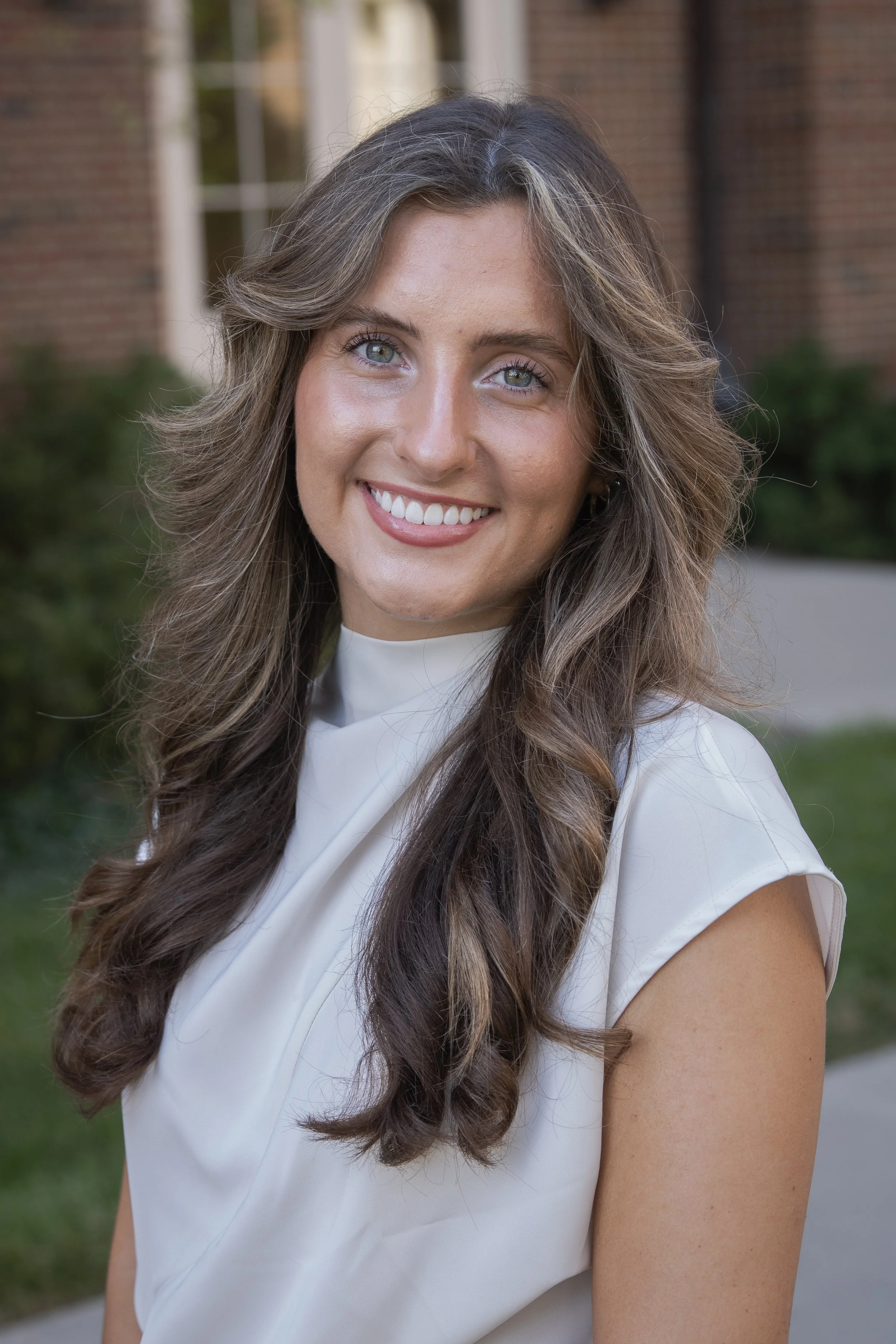 A smiling woman with long, wavy brown hair and light blue eyes, wearing a white top, standing outdoors with a brick building and greenery in the background.