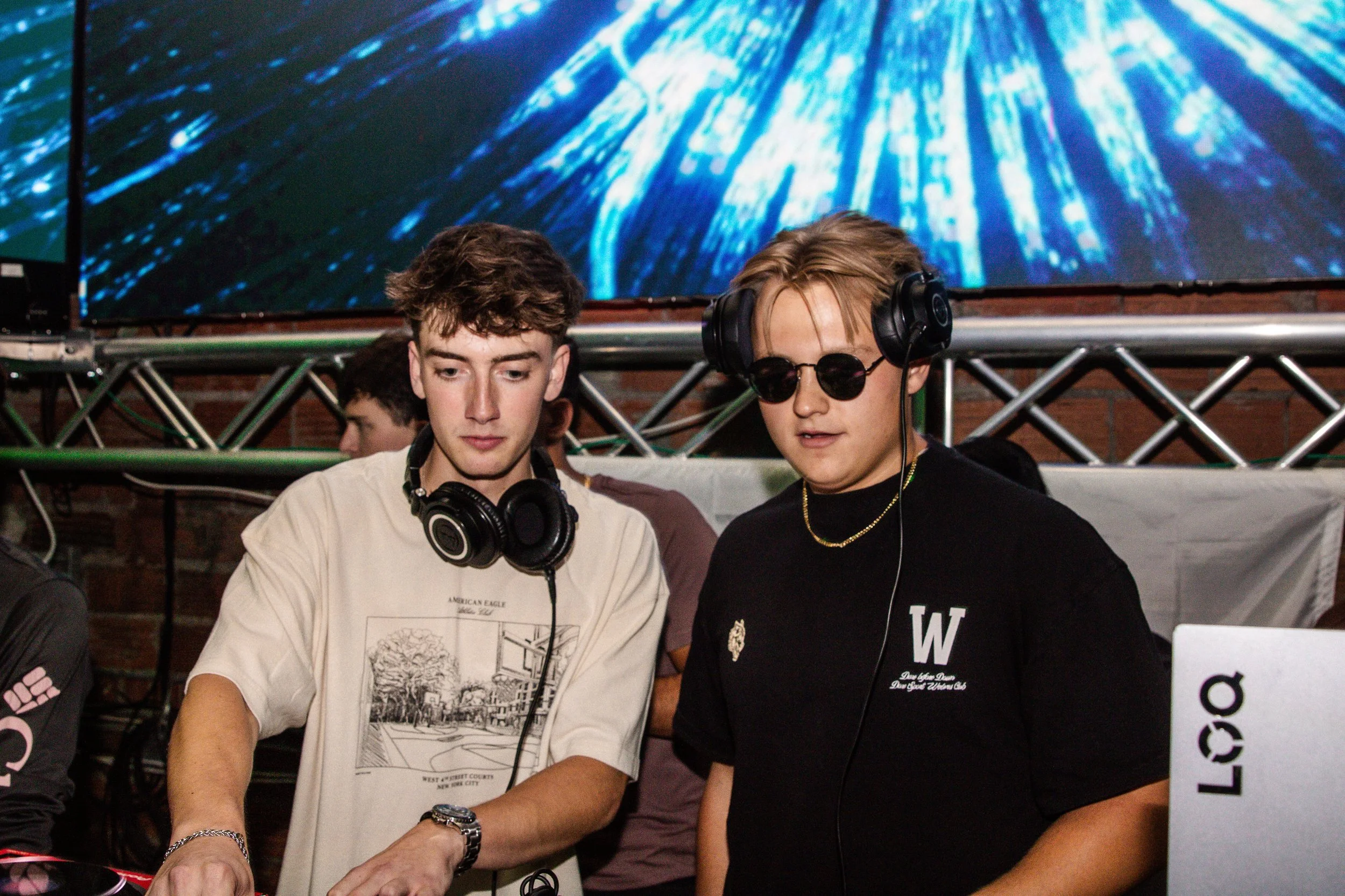 Two young men wearing headphones, one with sunglasses, stand in front of a DJ setup at a music event or party. A large digital screen with a blue abstract pattern is in the background.