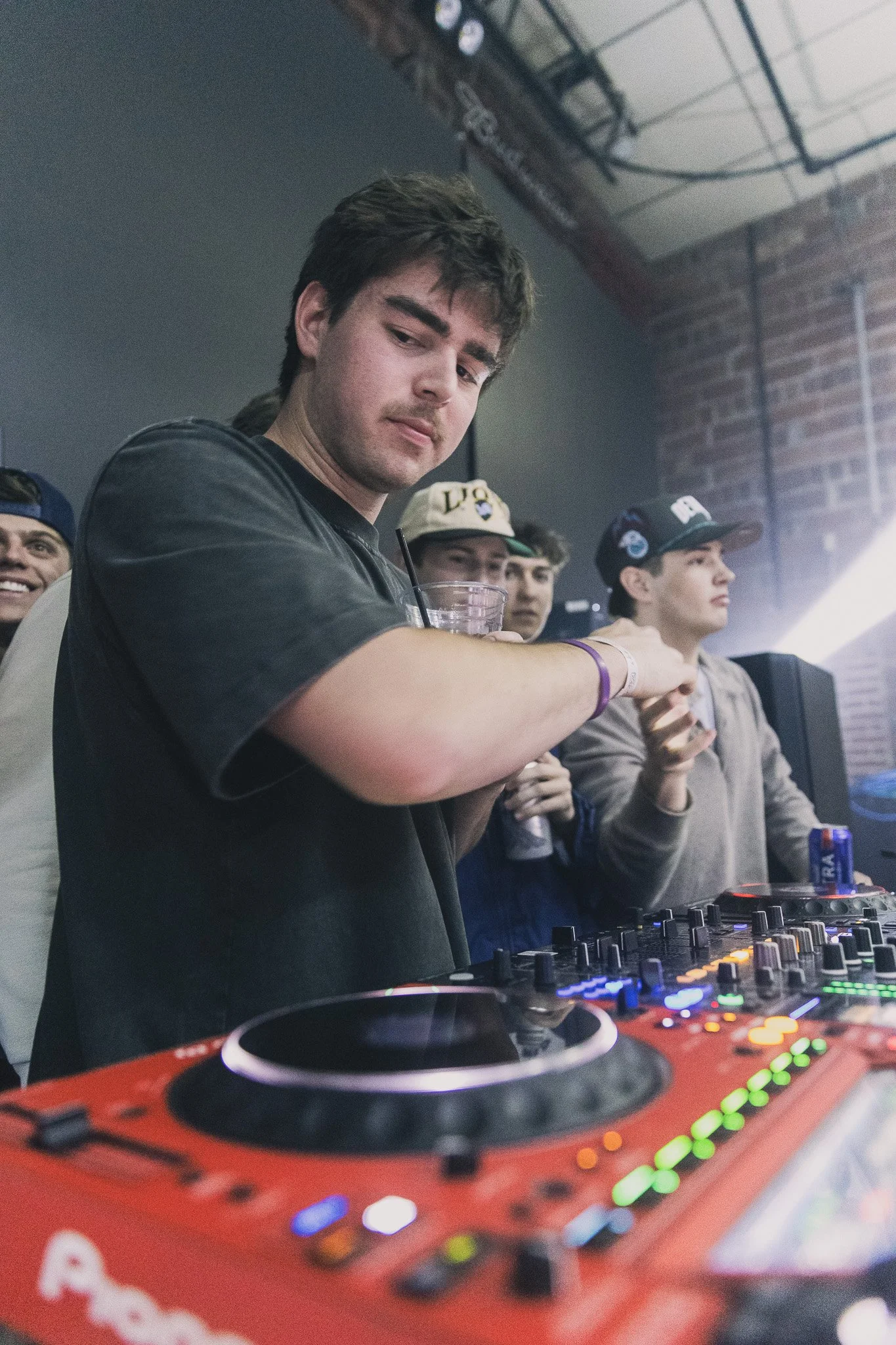 A young man at a DJ booth with other young men in the background, some holding drinks, in a club or concert setting.