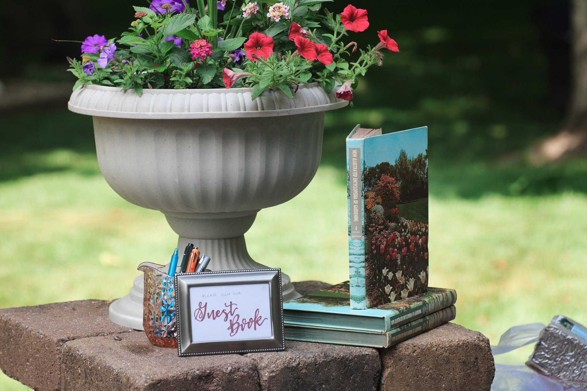A garden-themed sign-in book stand with alstroemeria flowers in a large white urn, a small framed sign that says 'Please sign our Guest Book,' and a few pens in a glass holder. A book with a picture of a garden scene is placed on stacked green hardco