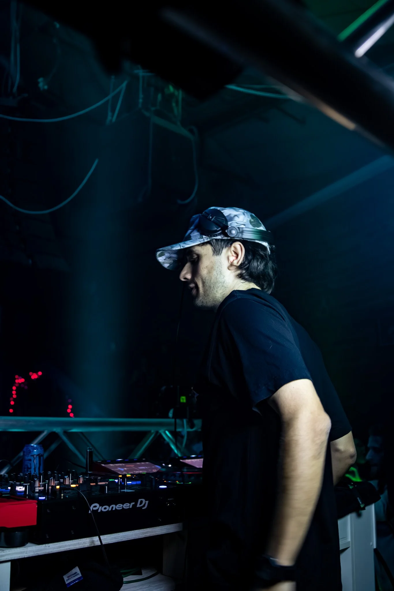 A male DJ wearing a camouflage cap and black t-shirt at a dimly lit nightclub.