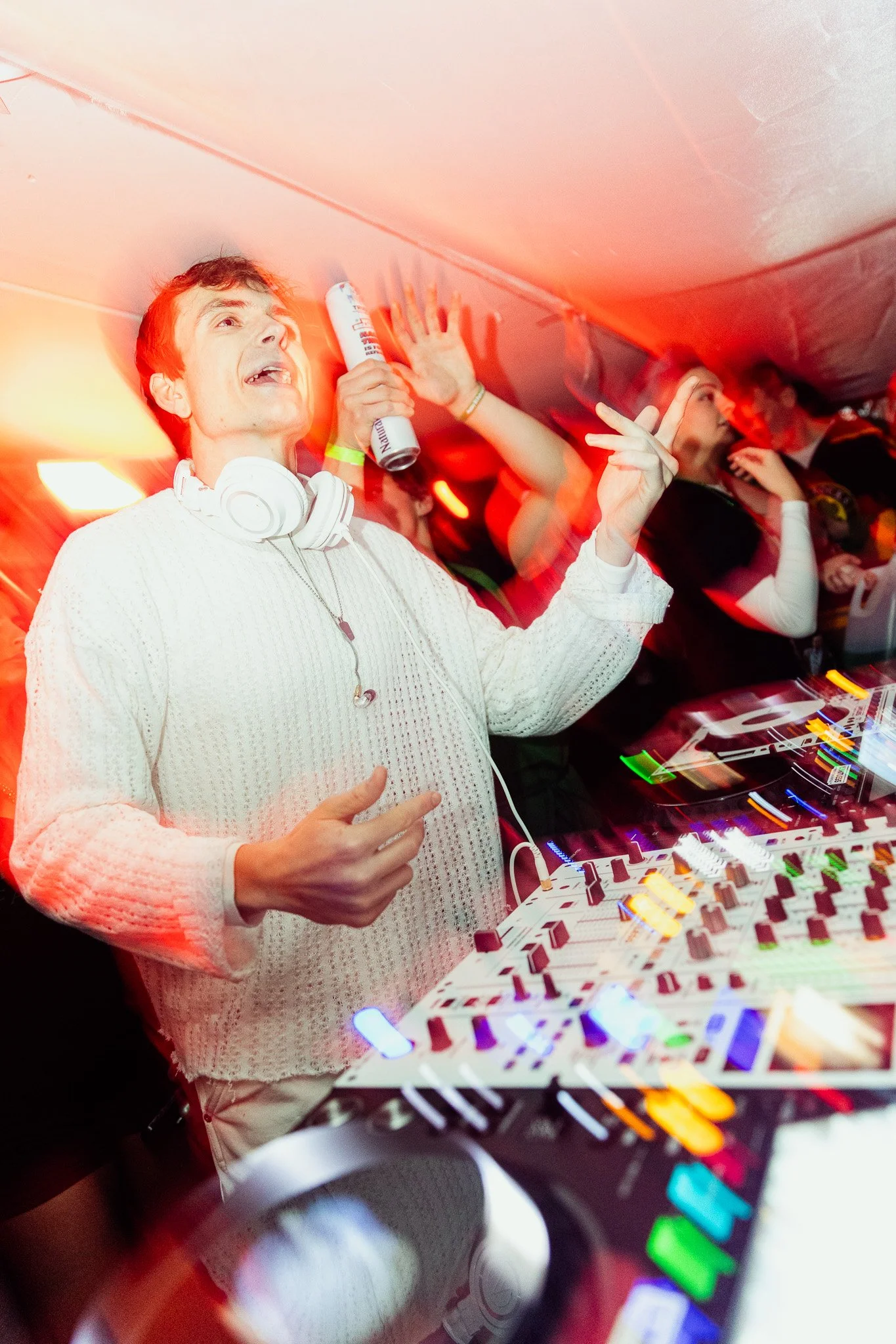 A DJ in a white sweater with headphones around his neck, holding a drink in one hand and making a peace sign with the other, at a lively party with colorful lights and a DJ setup.