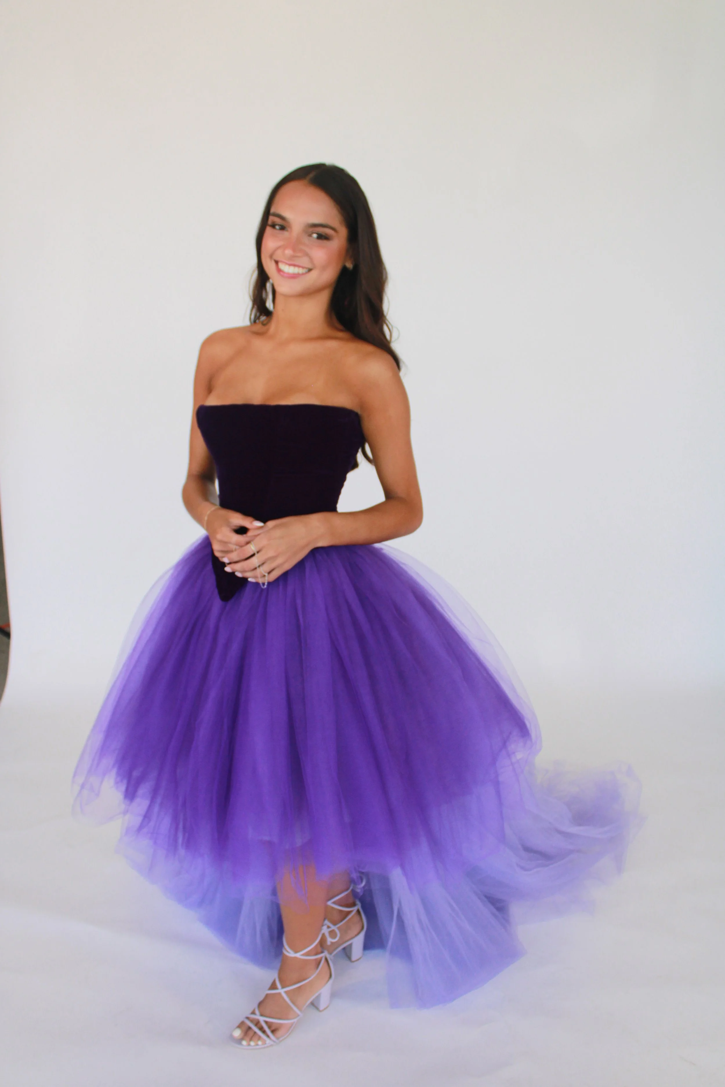 Young woman smiling in a black strapless top and purple tulle skirt, standing against a white background.