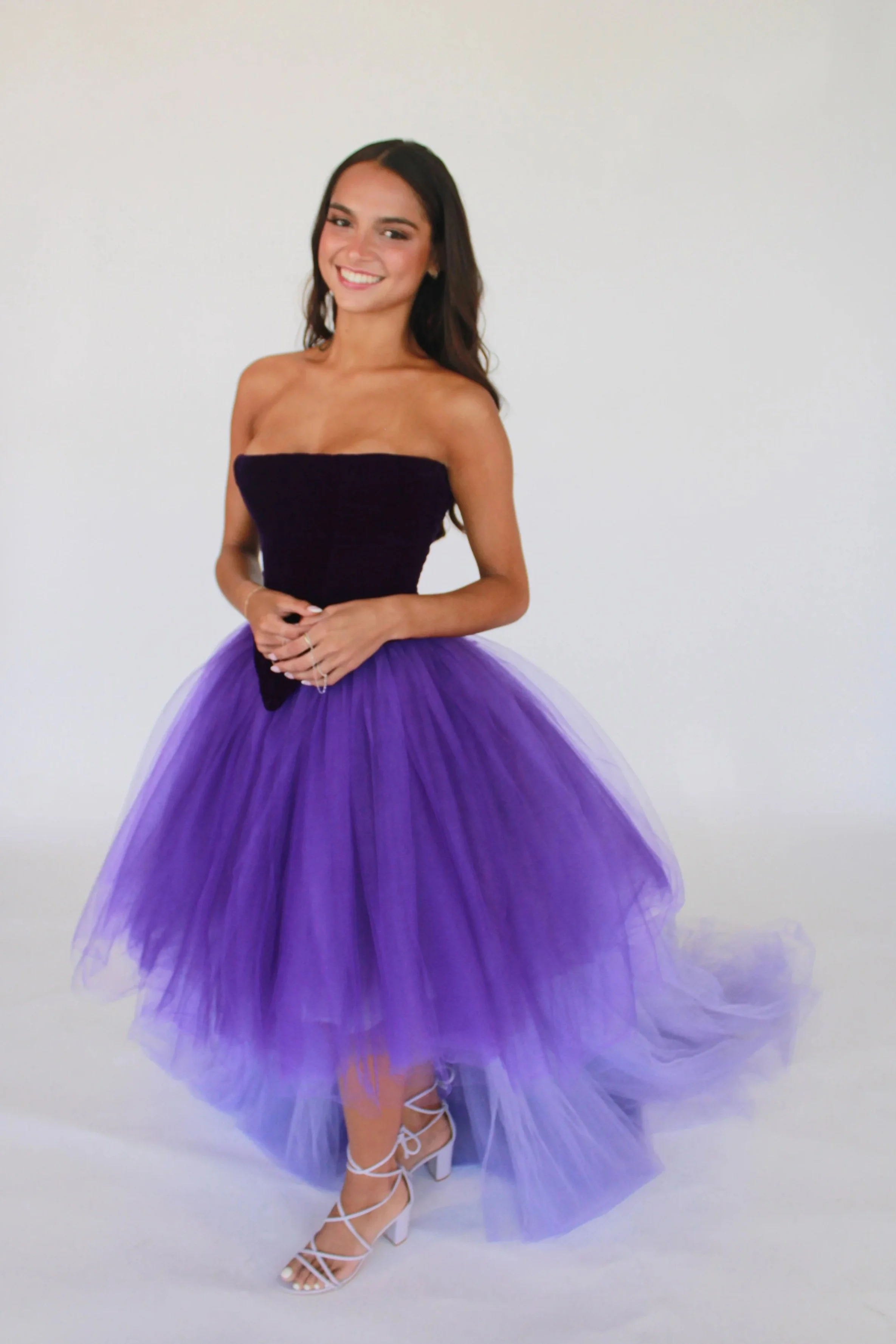 Young woman smiling, wearing a strapless black top and a purple tulle tutu skirt, standing against a plain white background.