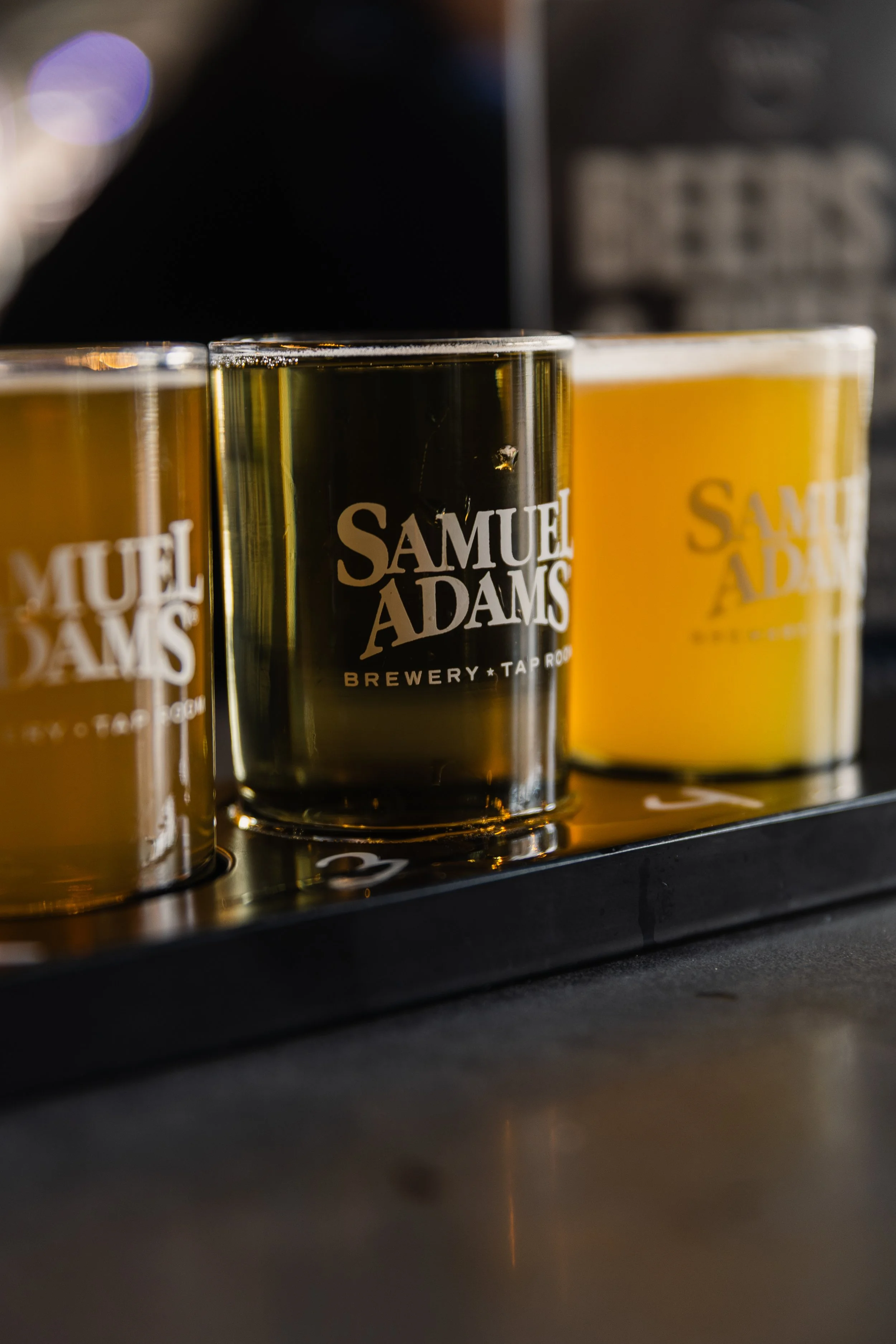 Three glasses filled with beer on a black tray, with the middle glass featuring a Samuel Adams logo, set against a blurred background.