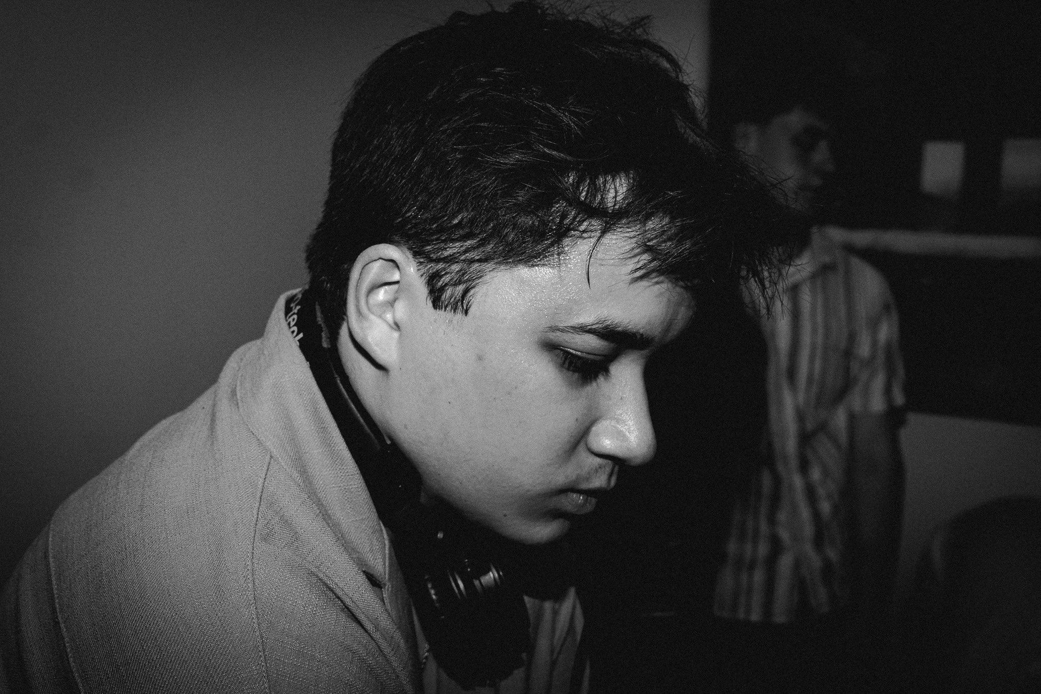 A young man with dark, short, messy hair and smooth skin, with his eyes closed or looking downward, wearing a light-colored jacket and headphones around his neck. Another person in a striped shirt is blurred in the background.