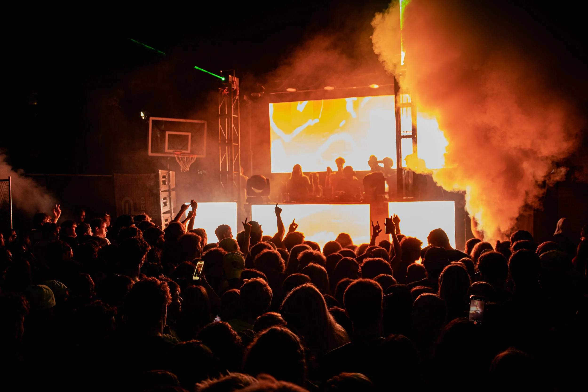 Crowd at a concert with a large stage, DJ booth, and bright LED screens showing abstract visuals, with fog and flames in the background.