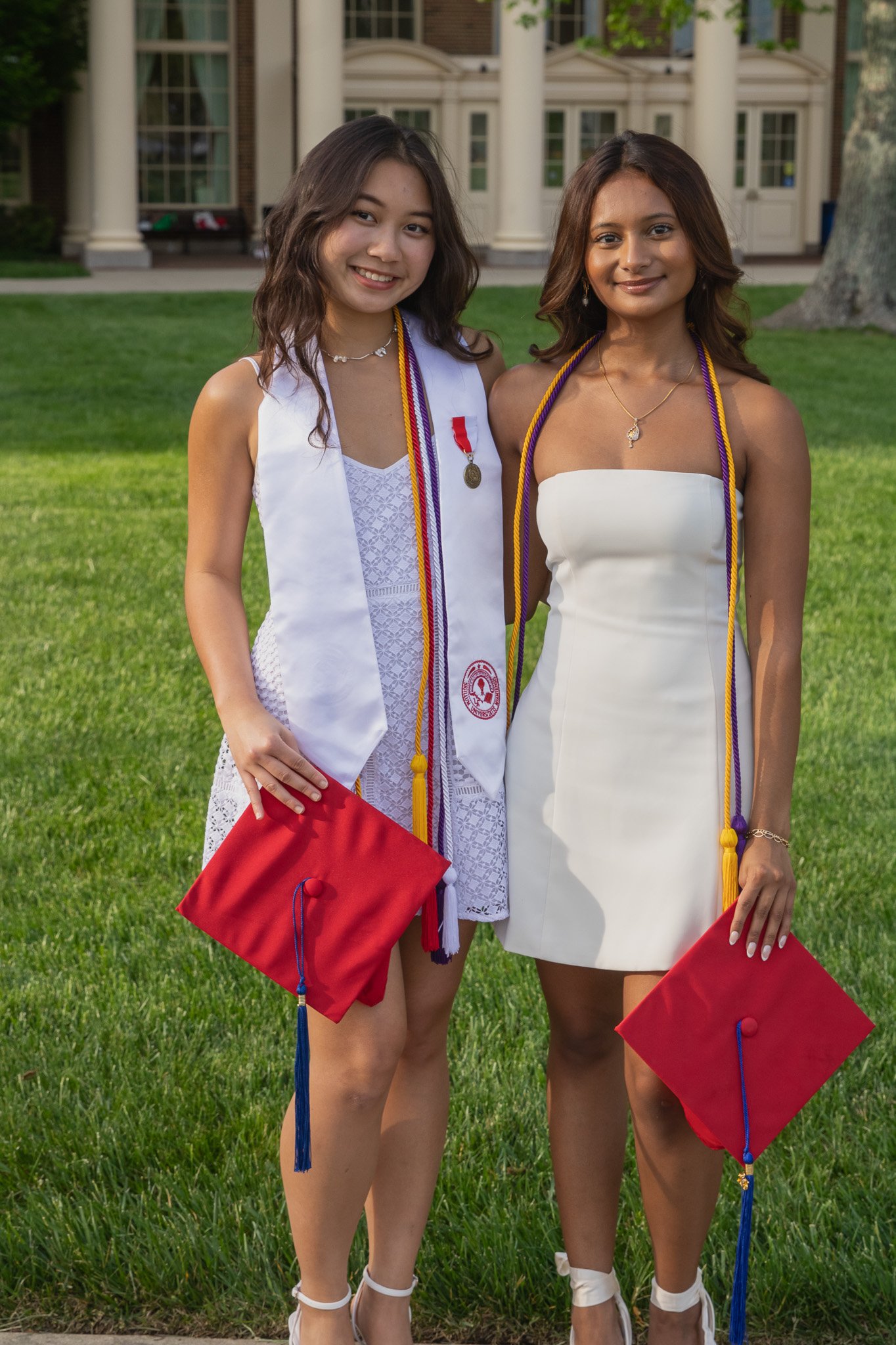 Two young women celebrating graduation outside on a green lawn. One wears a white dress with a white sash and holds a red graduation cap, the other in a strapless white dress with a red and white sash, both with medals and cords around their necks.