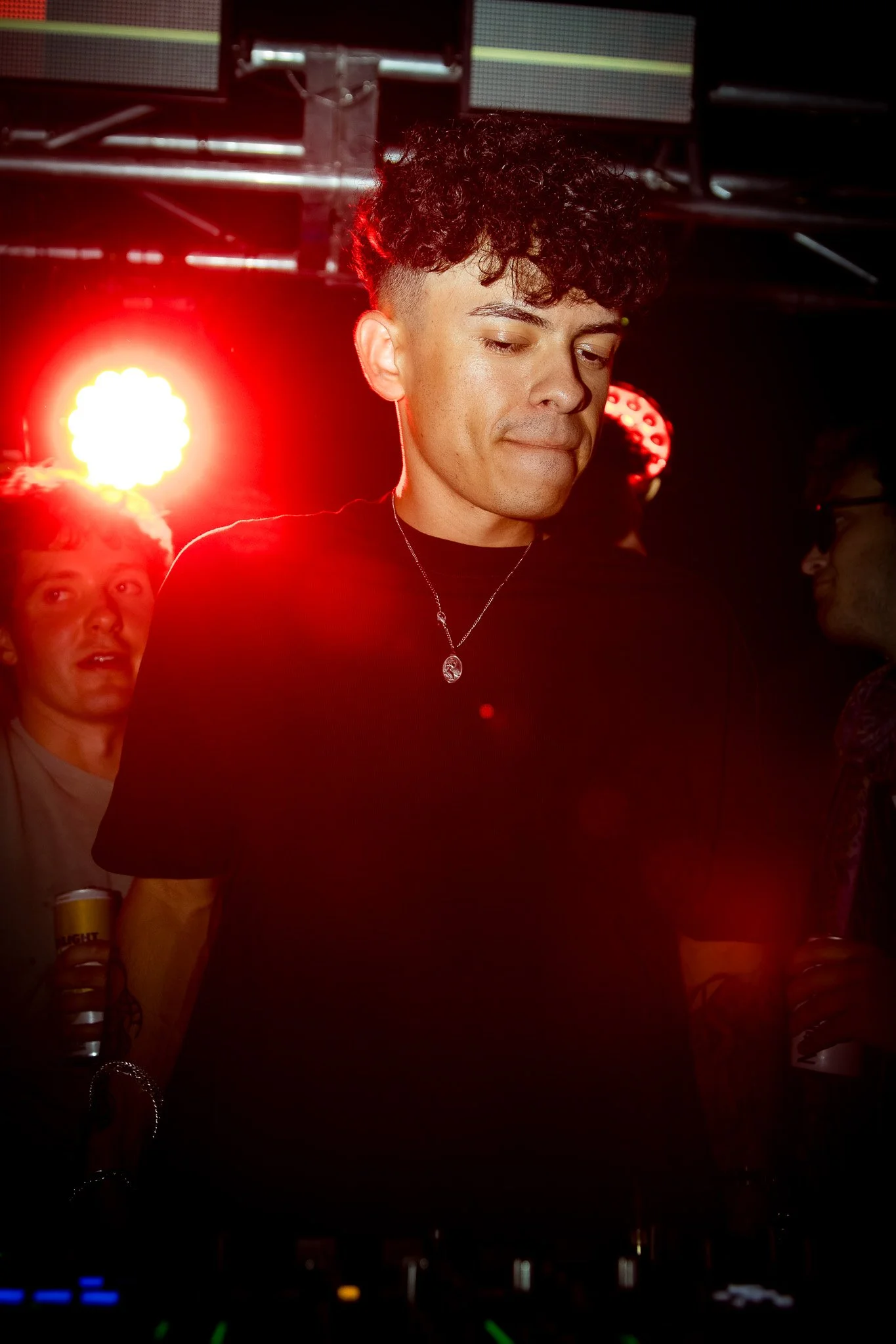 A young man with curly dark hair and light skin in a dark T-shirt, wearing a silver necklace with a pendant, standing in a dimly lit environment with red lighting and bright red and yellow lights behind him. Two people are visible in the background h