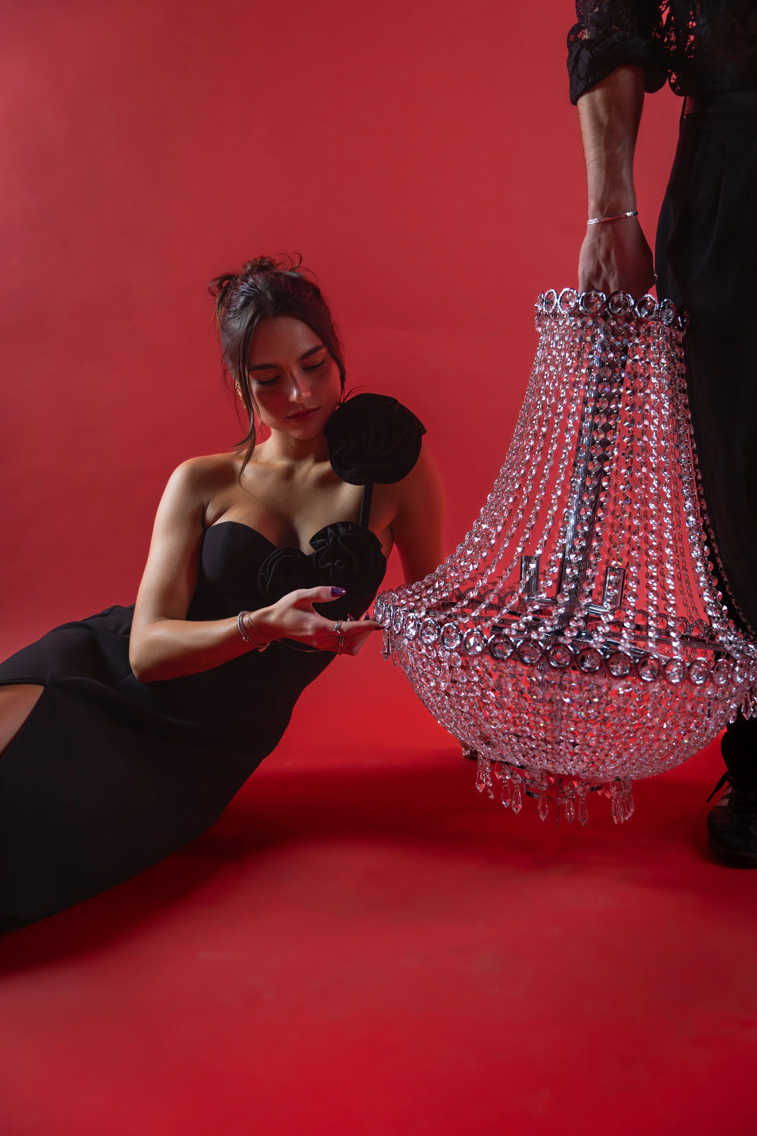 A woman dressed in a black strapless gown with floral embellishments on the bodice, kneeling on a red surface, looking at a large, ornate crystal chandelier being held by a man in black clothing. The background is red.