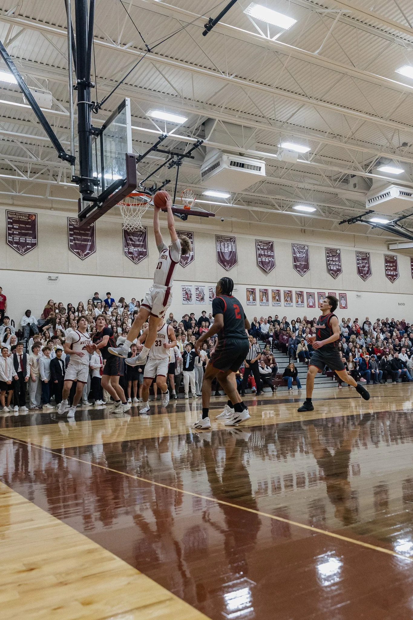 A basketball player in a white jersey is jumping to make a shot near the hoop, with other players and spectators watching in a gymnasium.