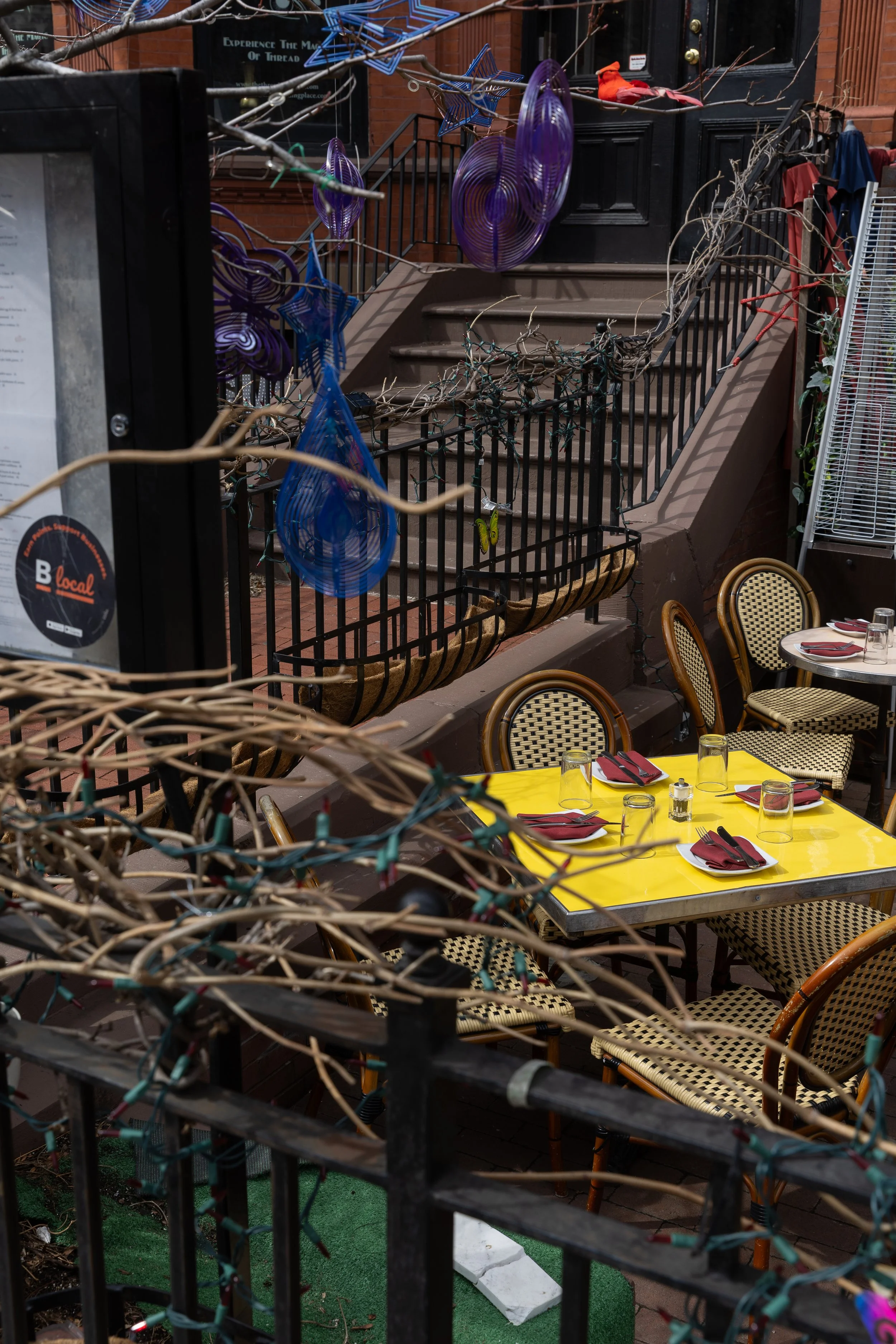 Outdoor restaurant patio with yellow table set for four, featuring glasses, utensils, and napkins, surrounded by woven chairs, decorated with strings of colorful birds and star-shaped ornaments hanging from a tree branch, and a railing with twinkling