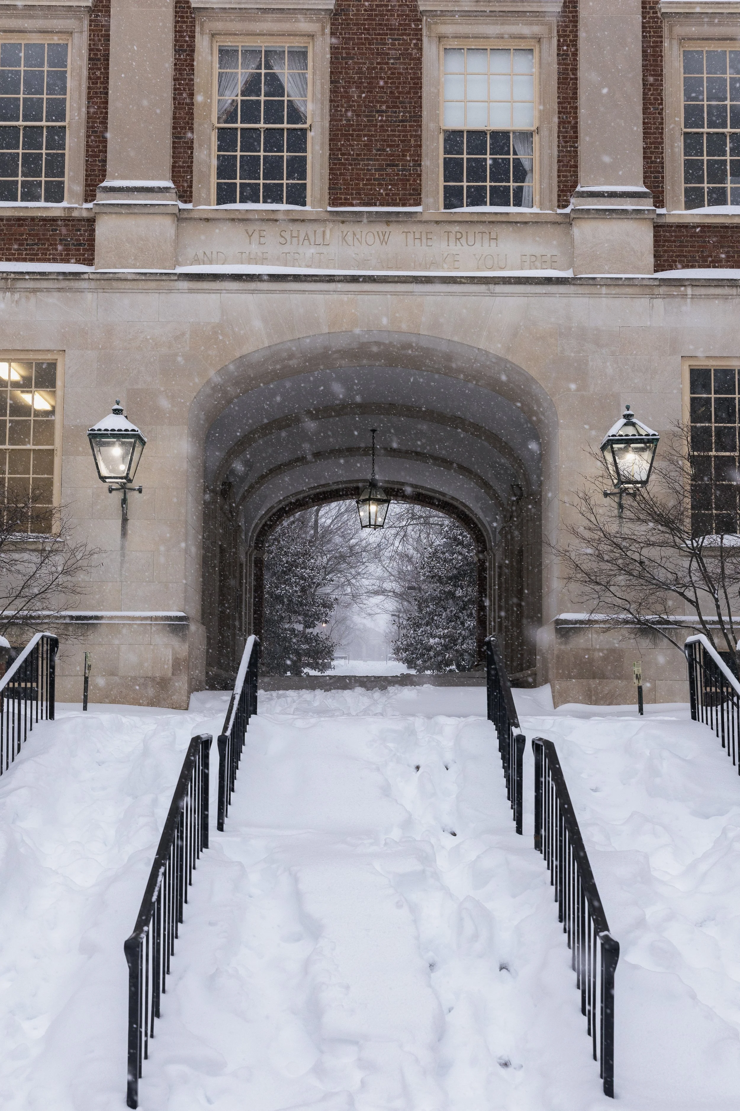 Snow-covered entrance with an arched doorway, two lamp posts, and footprints in the snow leading inside a brick building, with a quote engraved above the arch reading 'Ye shall know the truth and the truth shall make you free.'
