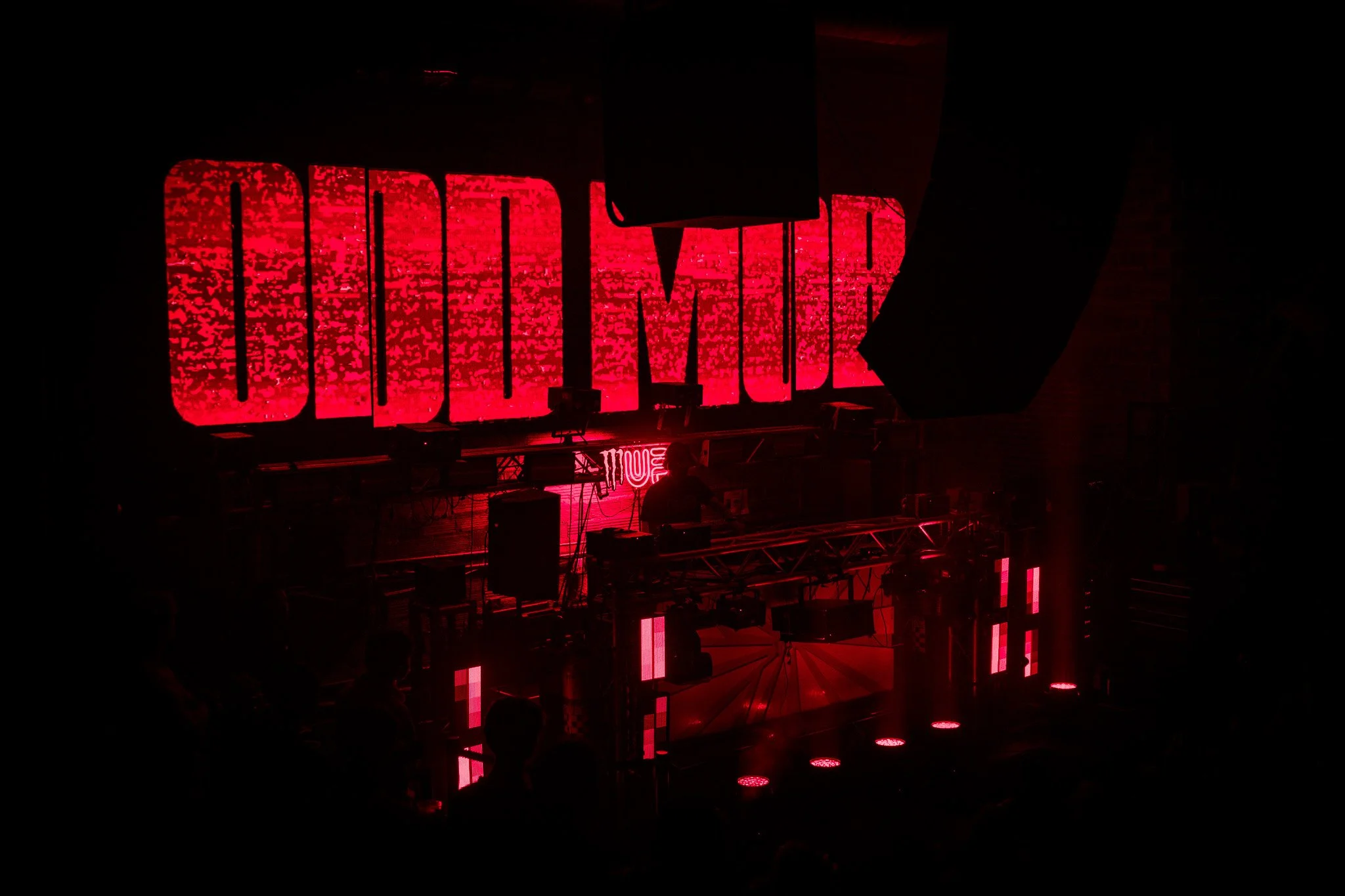 Red neon sign reading 'DOOM' on a dark stage with DJ equipment and silhouettes of people.