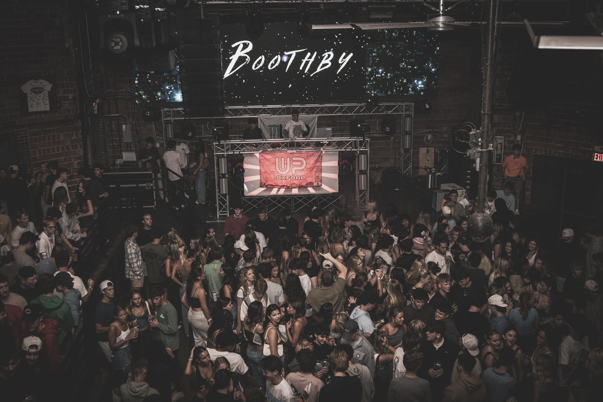 Crowd of young people dancing and socializing at a nightclub or concert venue with a DJ on stage and a large screen that says 'Bootby' in the background.