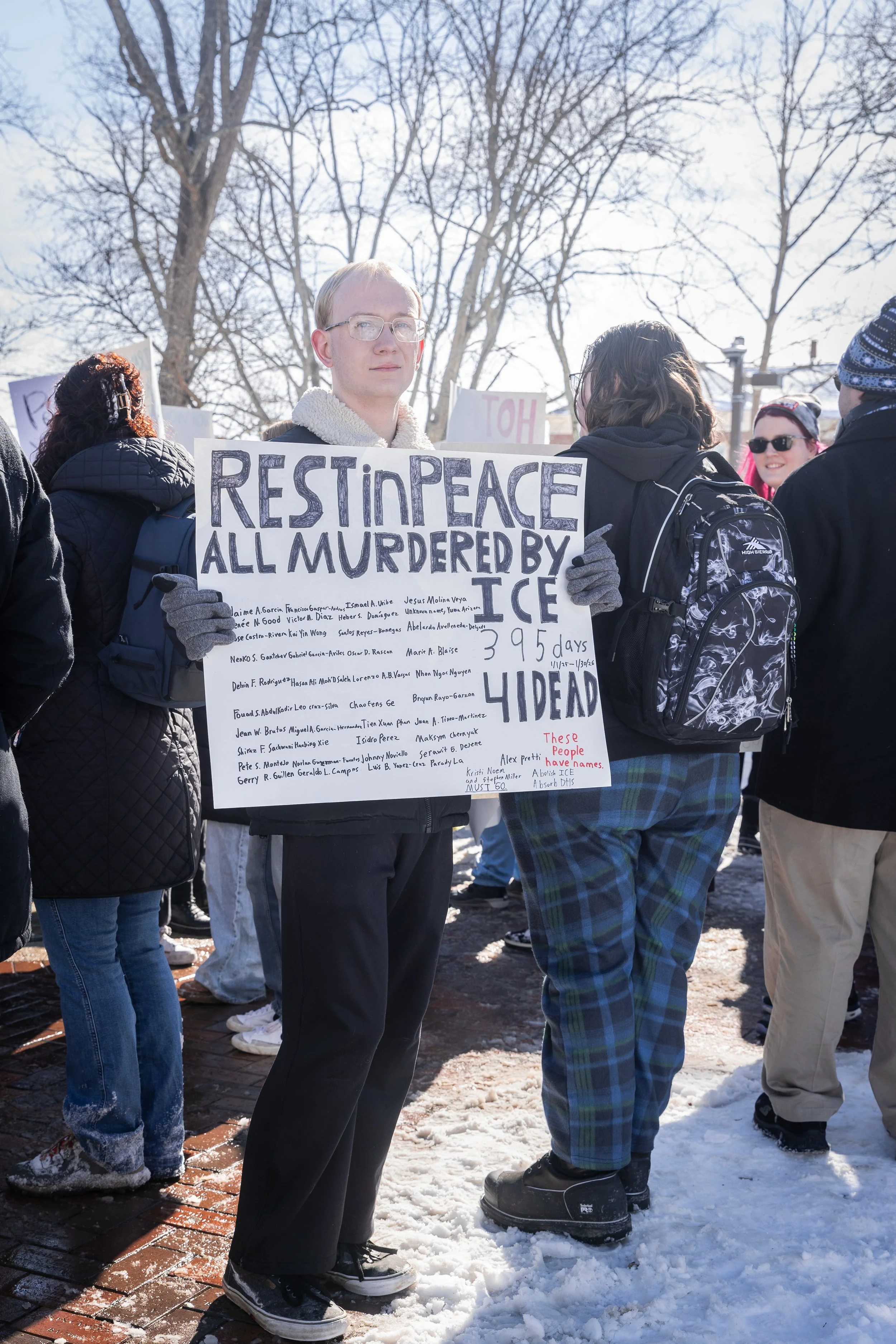 A young man wearing glasses, gloves, and a black jacket holds a sign that reads, 'Rest In Peace All Murdered By ICE,' during a protest in winter. Several other people are gathered near him, some wearing backpacks, and there are leafless trees in the 