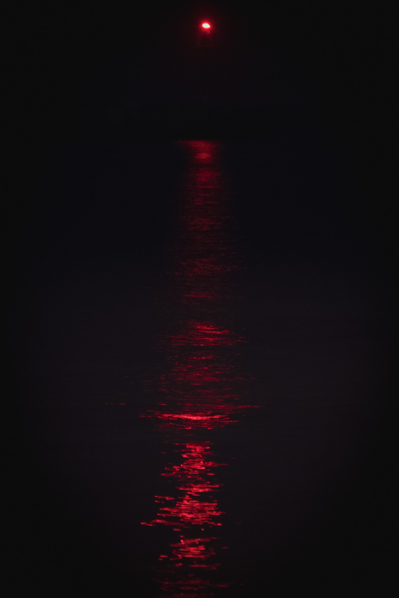 A dark scene with a single red light at the top and its reflection shimmering on the water below.