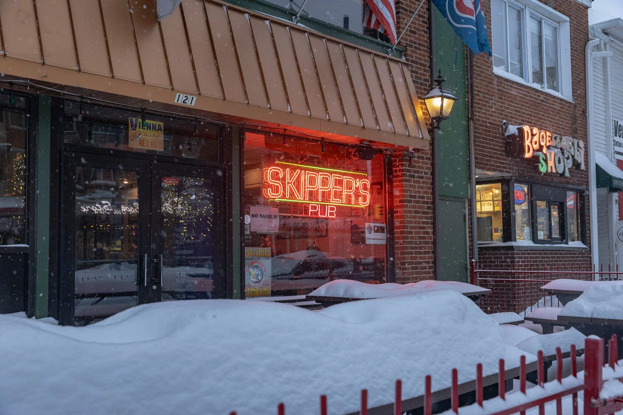 Snow-covered sidewalk in front of a bar with a neon sign that reads 'SKIPPER'S PUB' and another sign that reads 'BAGEL SHOP' on the building.