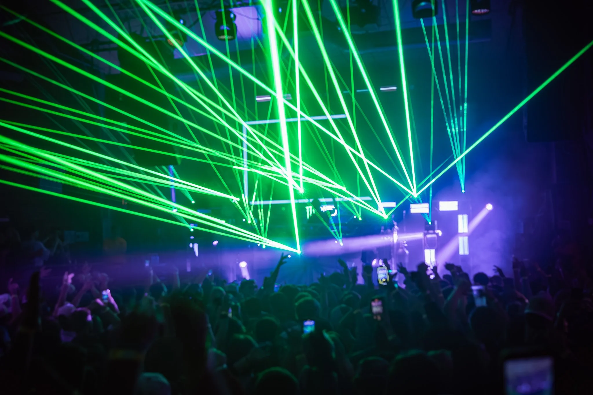 A concert or nightclub scene with green laser lights crisscrossing above audience members, some of whom are taking photos or recording with their phones.