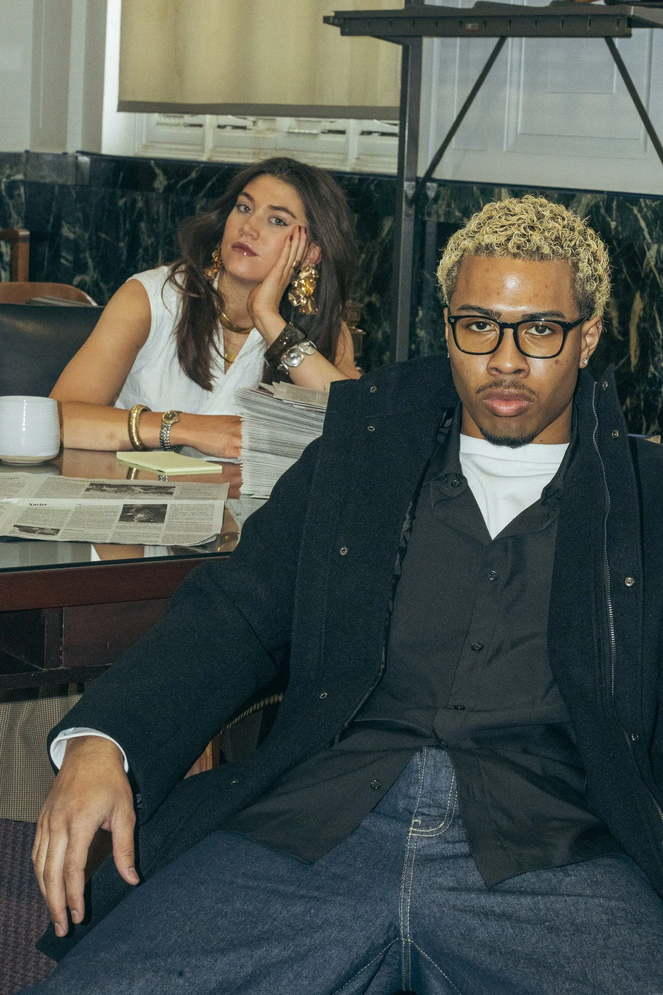 A young man with glasses and curly blonde hair sitting in the foreground with a serious expression, wearing a black jacket, black shirt, and jeans. Behind him, a woman with long dark hair, wearing large gold earrings, a white sleeveless top, and vari