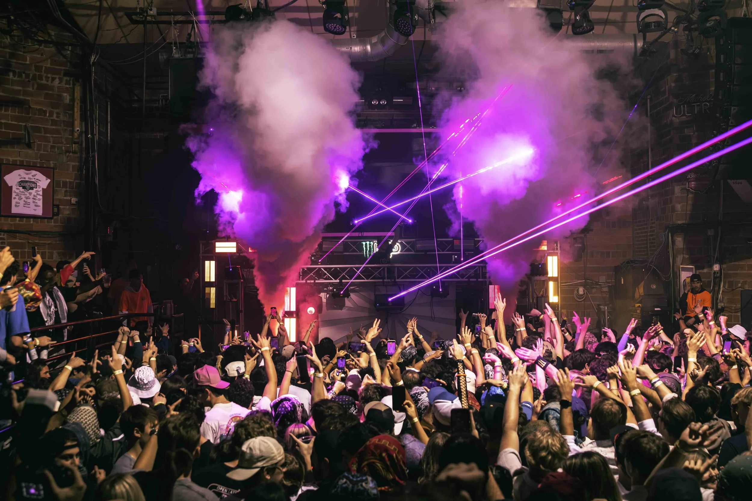 Crowd of people at a concert with colorful laser lights, pink and purple smoke effects, and a DJ on stage.