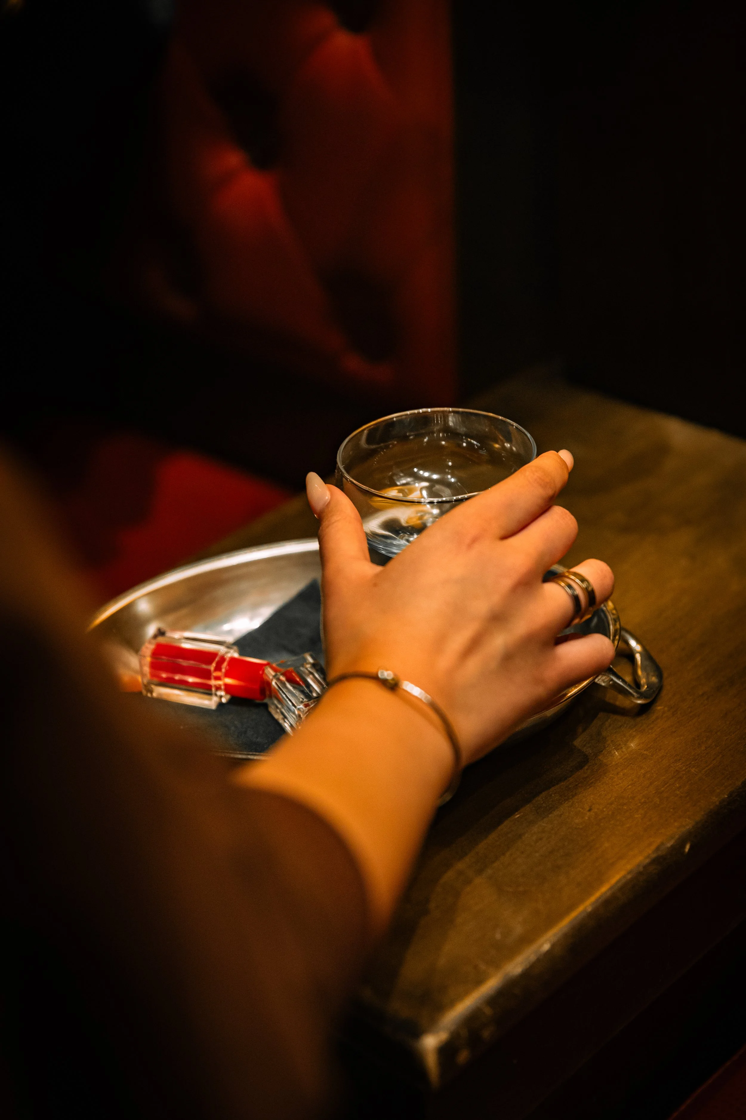 A person holding a glass of water on a tray, with a safety pin and makeup lipstick on the tray, in a dimly lit setting.
