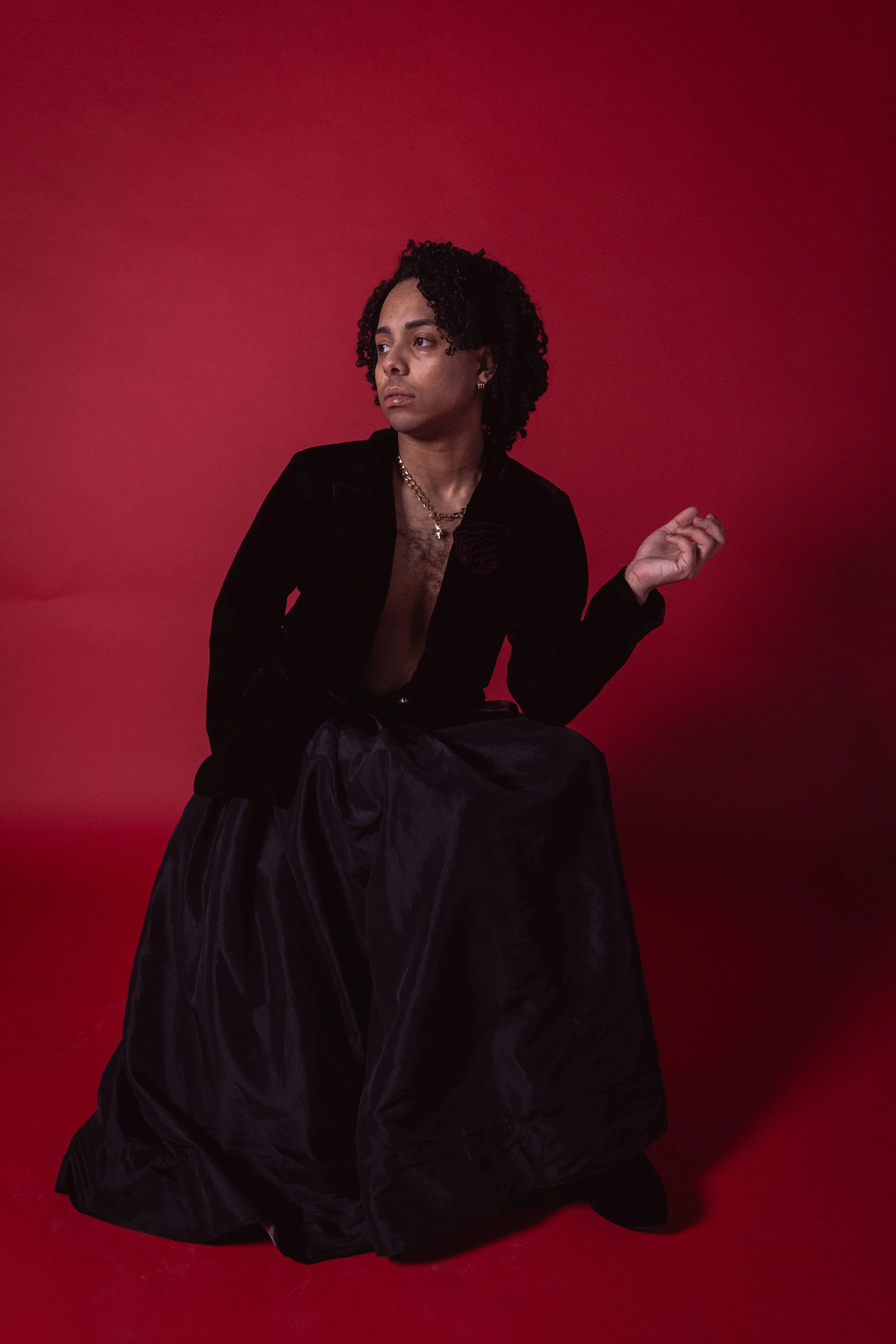 A person with dark curly hair sitting against a red gradient background, dressed in a black velvet jacket and long black skirt, looking to the side.
