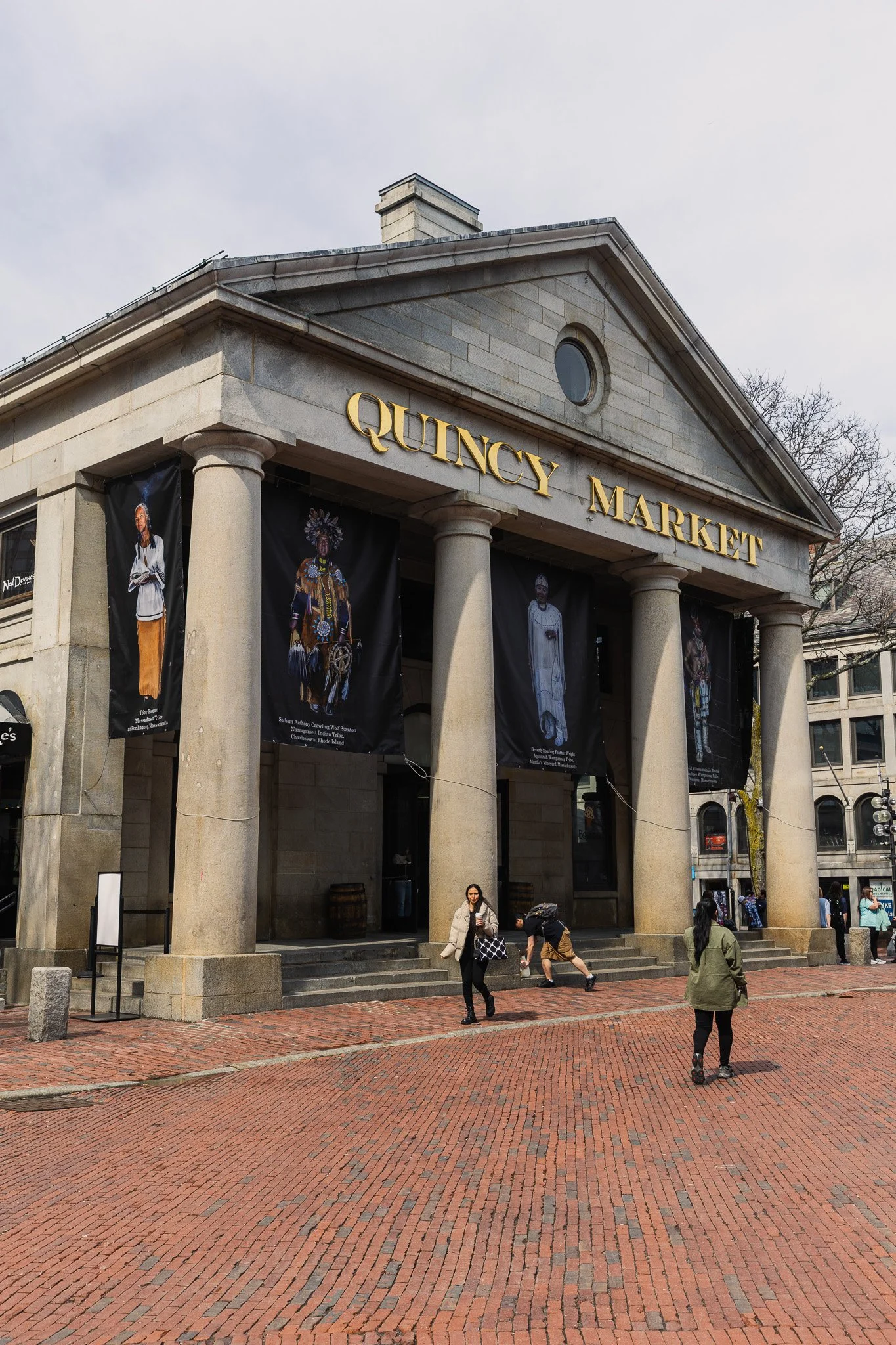 Exterior of Quincy Market building with large columns and banners displaying artwork and people, with pedestrians walking by.