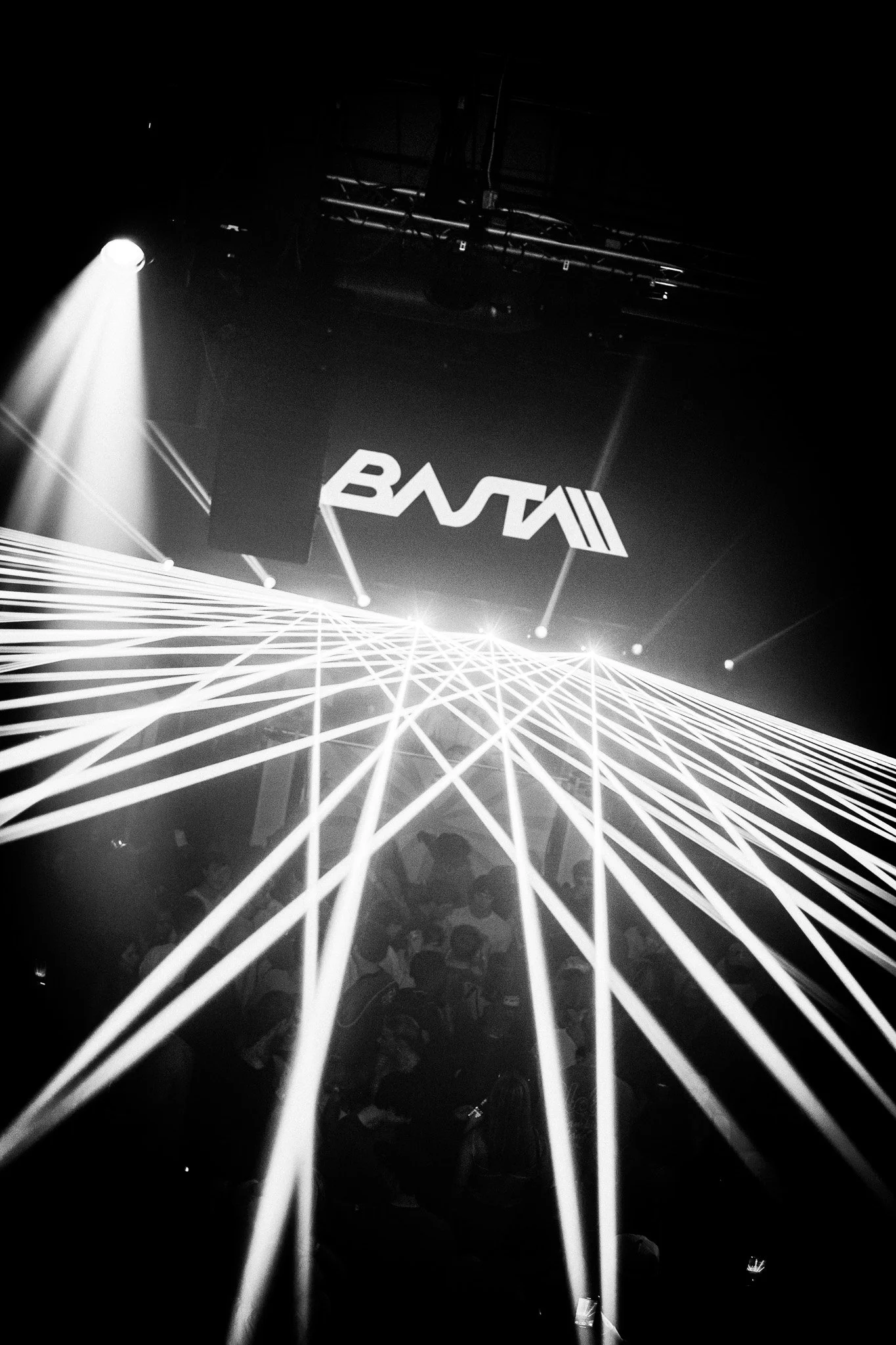 A black and white photo of a concert or event scene with laser lights projecting geometric patterns, a large screen displaying the BNT logo, and a crowd of people.