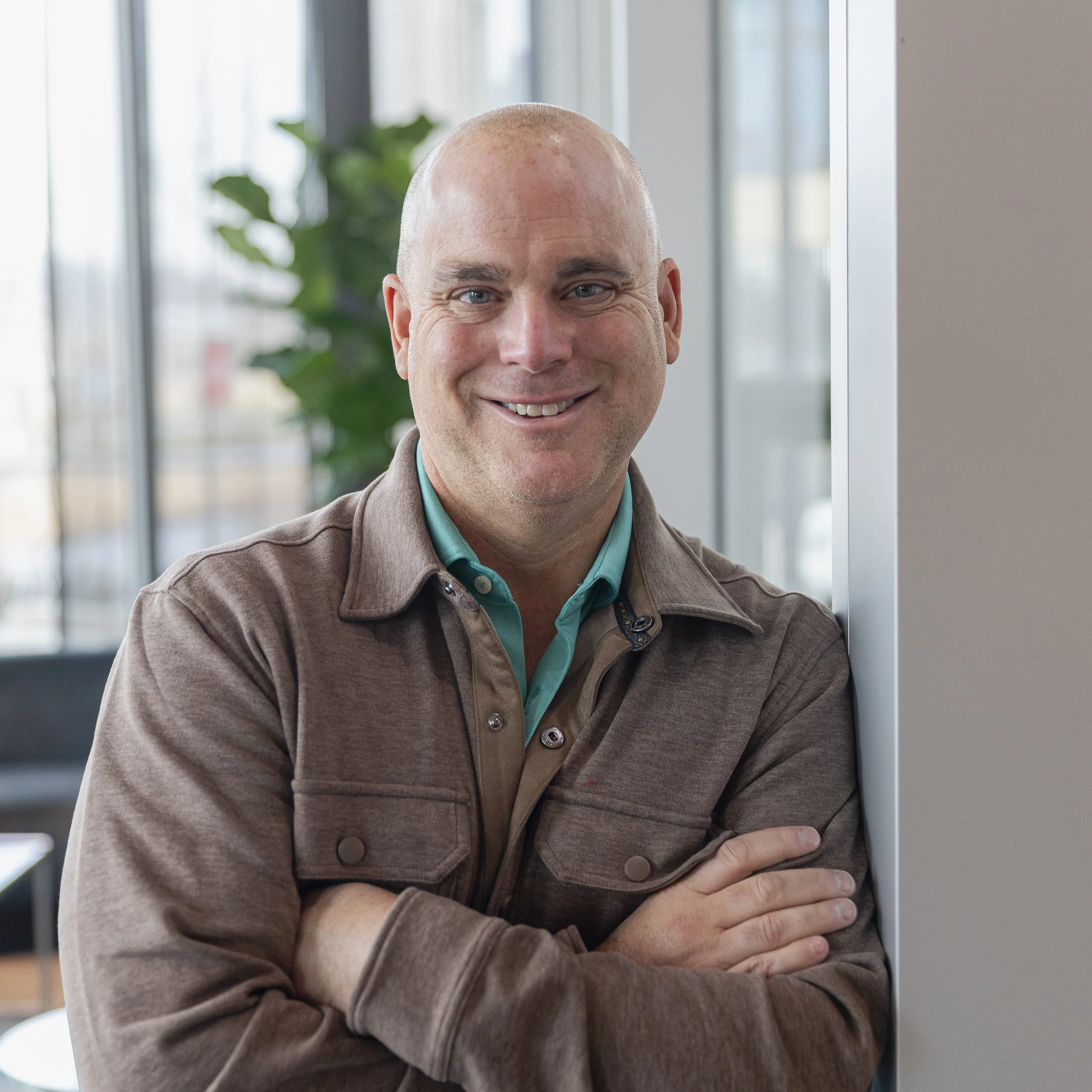 A smiling man with a shaved head standing indoors near a window with a view of a building and trees outside, wearing a brown jacket and a teal shirt.
