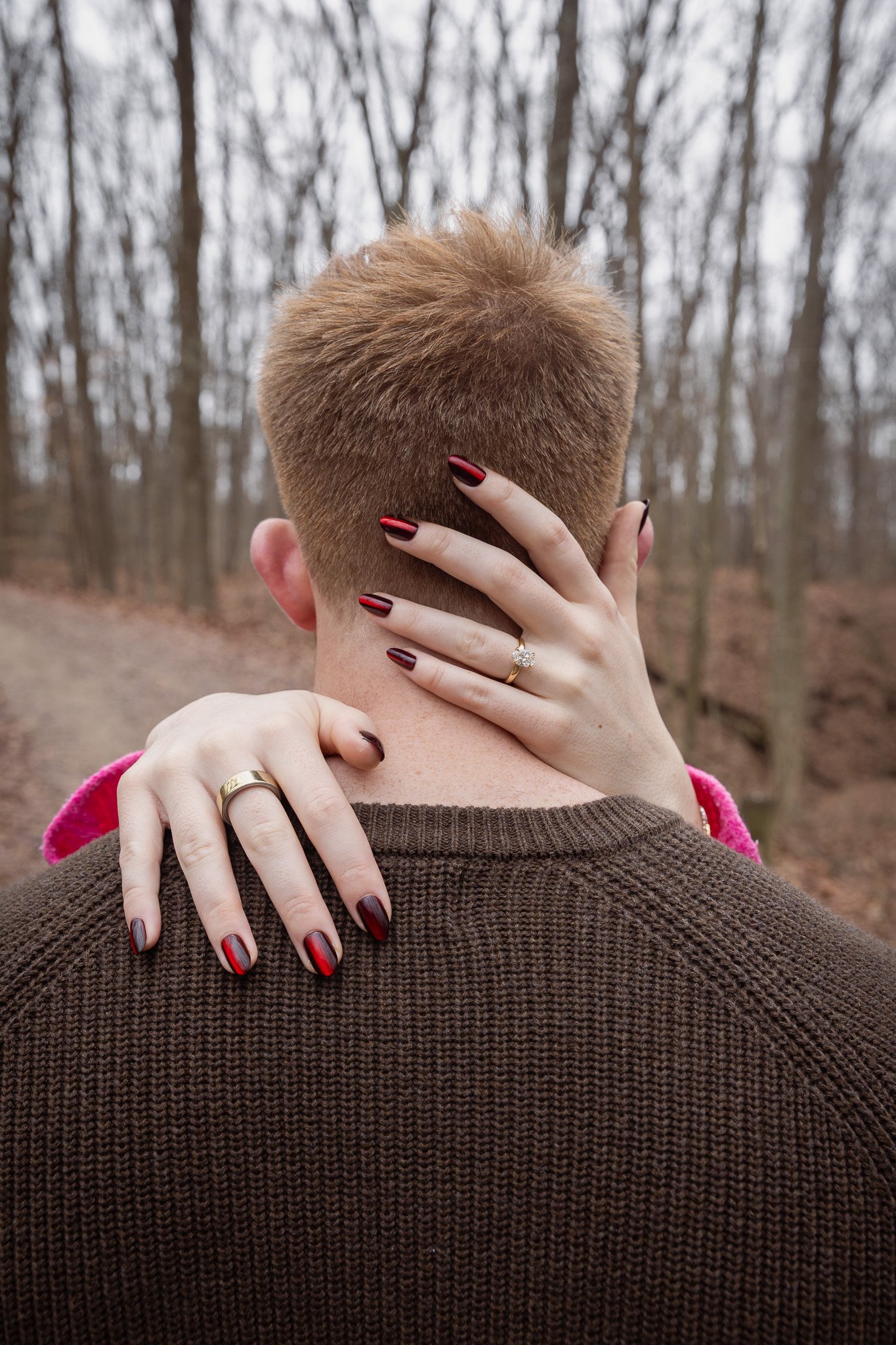 A woman and a man stand in a wooded area, with the woman wrapping her arms around the man's neck, revealing her ring and painted nails, while he is facing away from the camera.