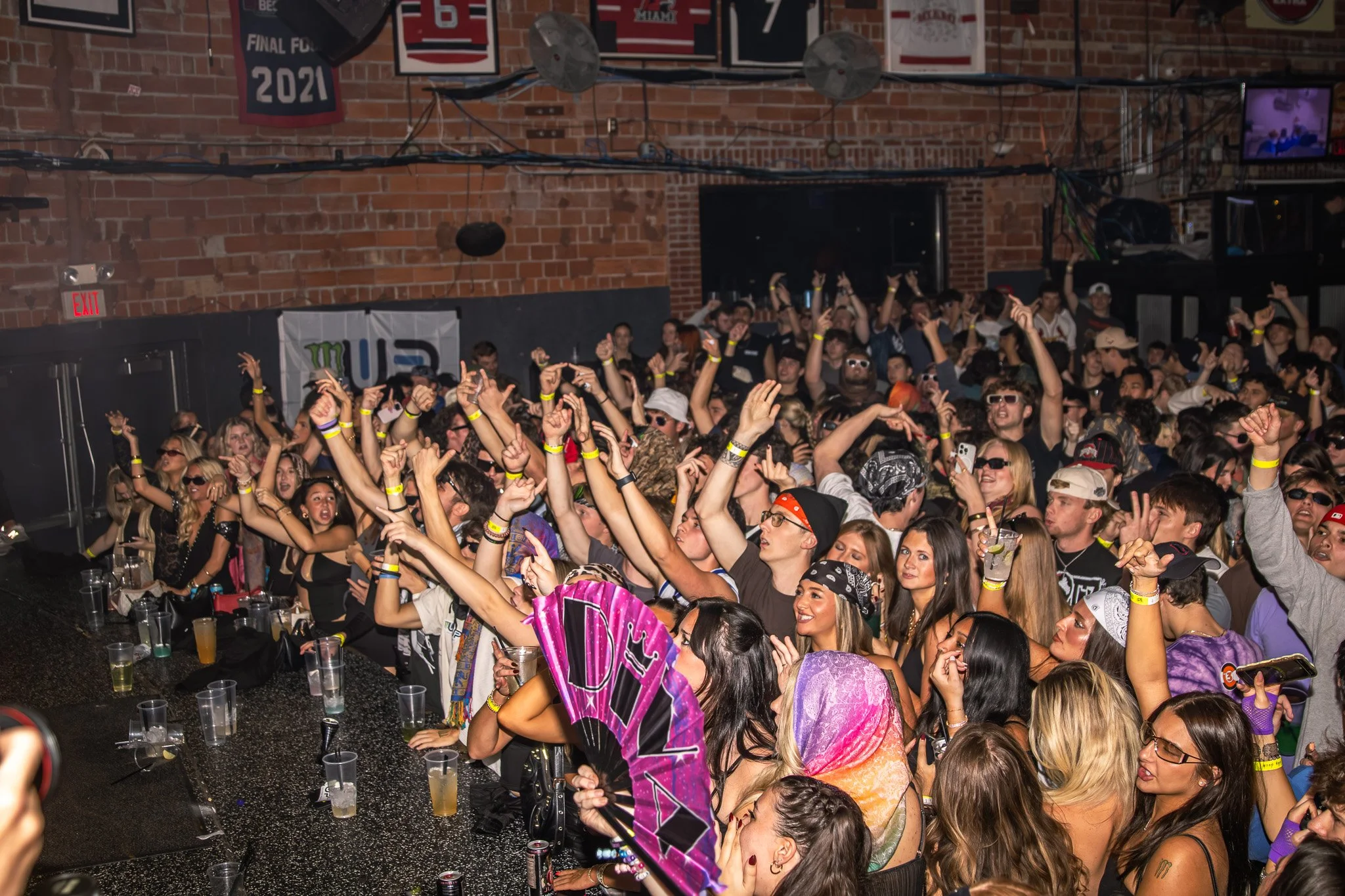 A large crowd of people at a concert or club, dancing and enjoying music, with some holding drinks and taking photos, in a brick-walled venue with banners and screens.