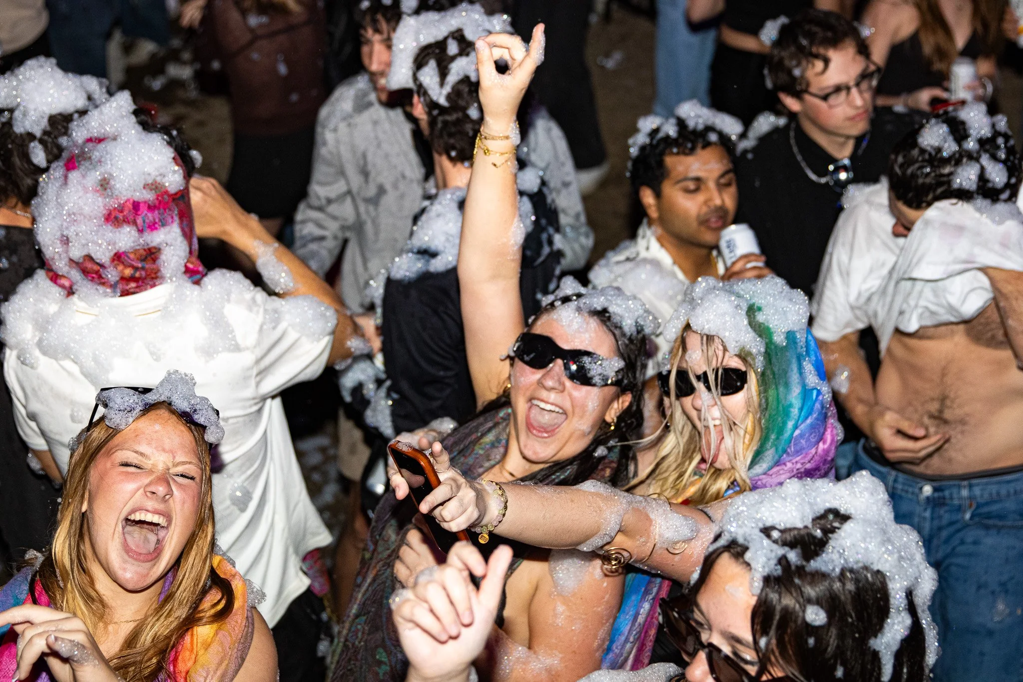 People celebrating at a foam party with soapy bubbles, some wearing sunglasses and colorful rain ponchos, dancing and having fun.