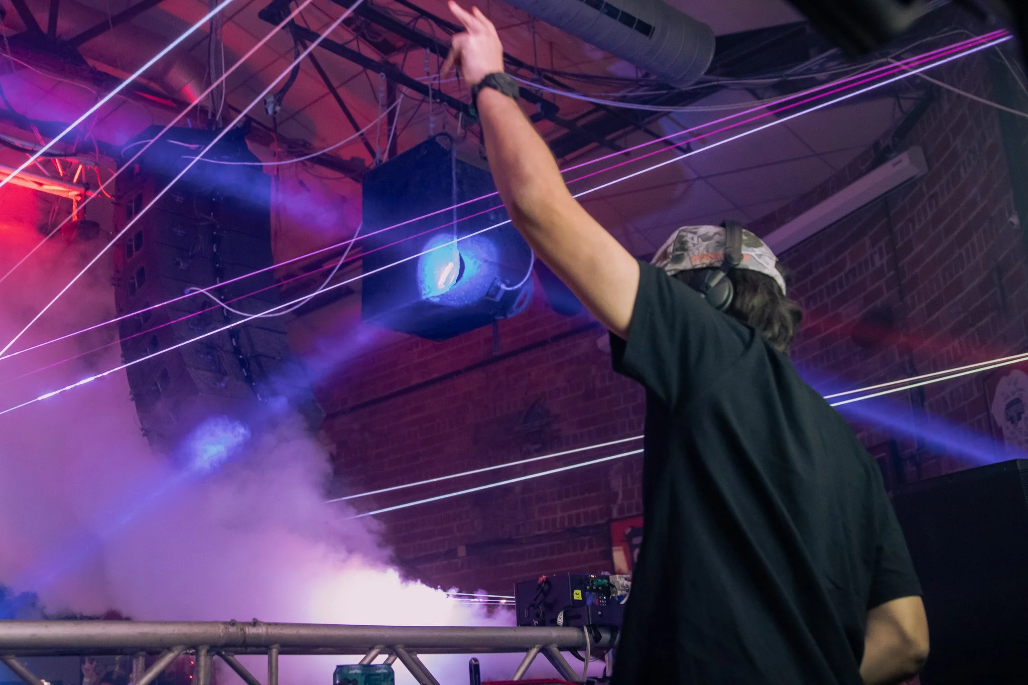 A DJ wearing headphones and a cap is seen at a nightclub or concert, with colorful laser lights and smoke effects creating a lively atmosphere.