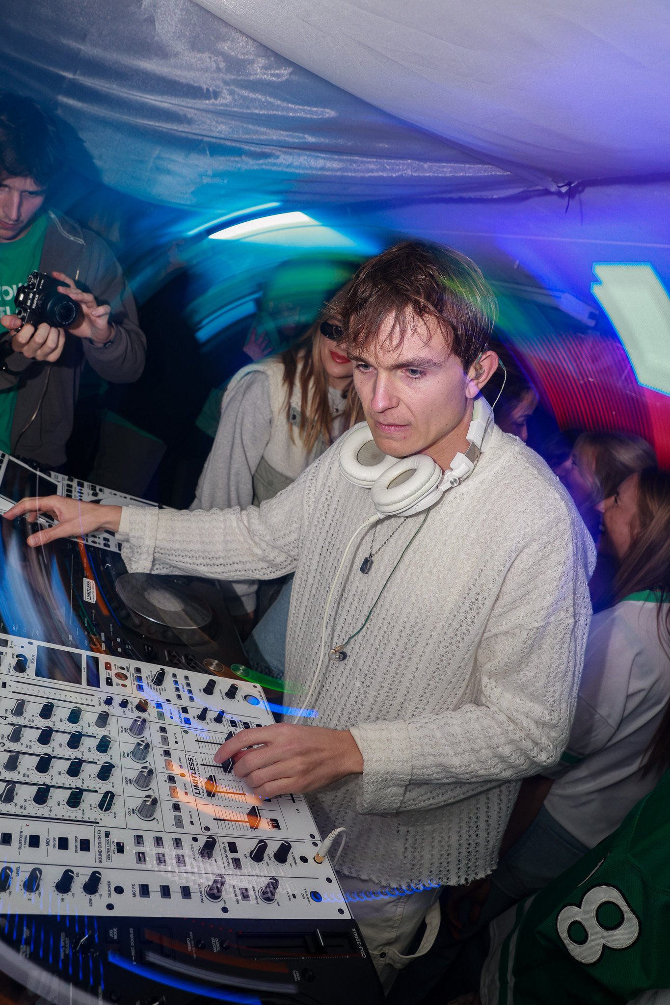 A young DJ with headphones around his neck operating a mixing console at a party, with other people in the background and colorful lighting.