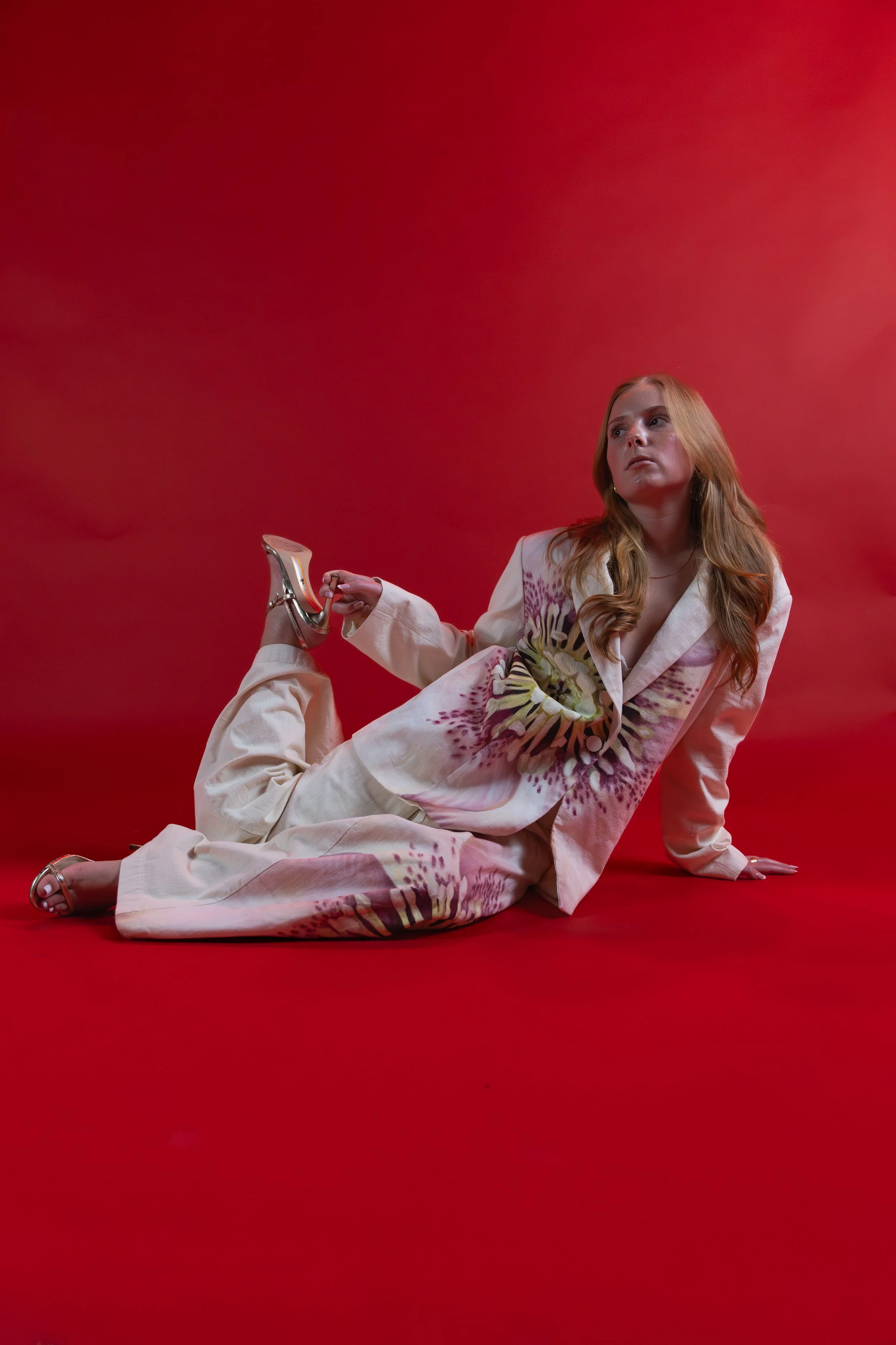 A woman with long red hair, wearing a floral patterned cream-colored suit and high heels, is posed reclining on a red background, looking off to the side.