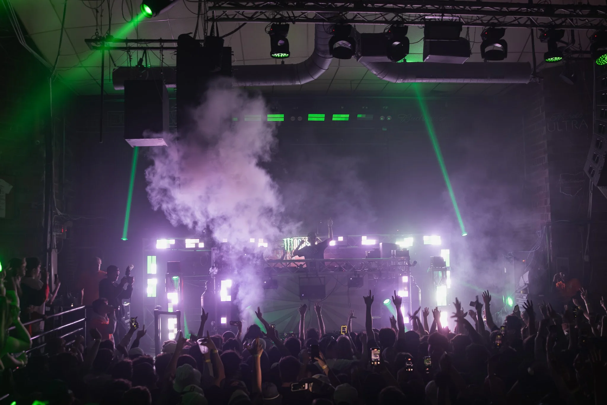 A live DJ performs on stage at a crowded concert with green laser lights, fog effects, and a cheering audience taking photos.