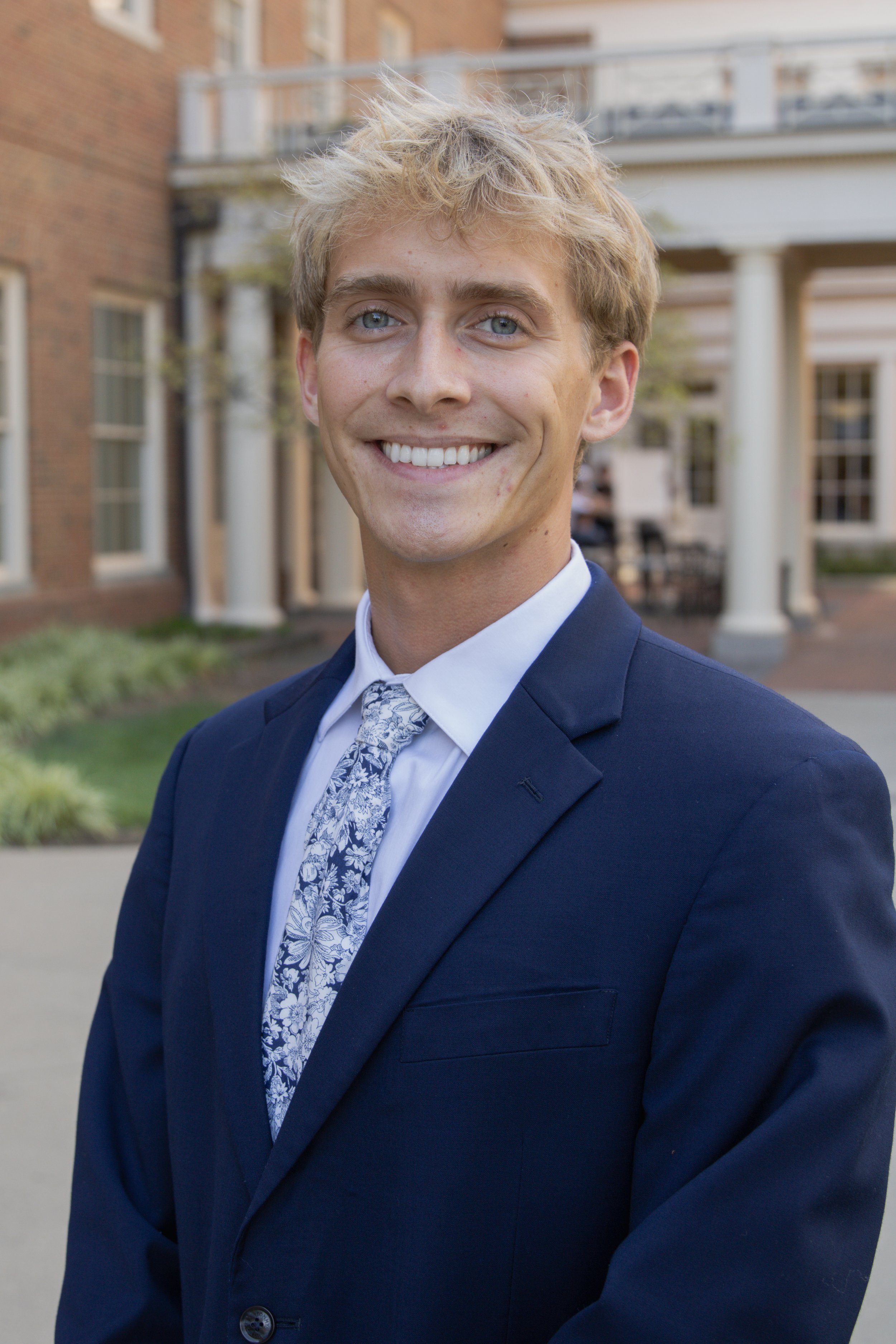 A young man in a navy suit with a floral patterned tie, smiling outdoors in front of a brick building with white columns.