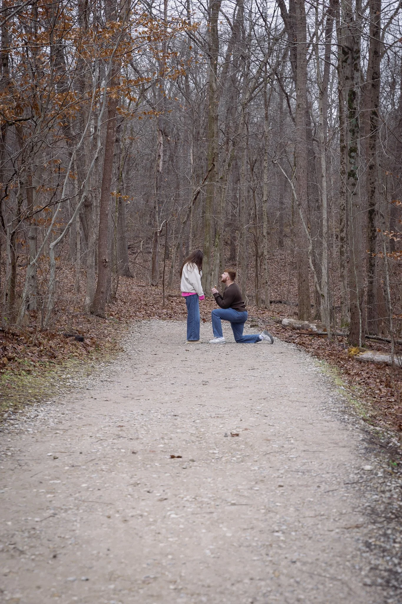 A man kneeling on one knee on a dirt trail in a forest, proposing to a woman standing in front of him.