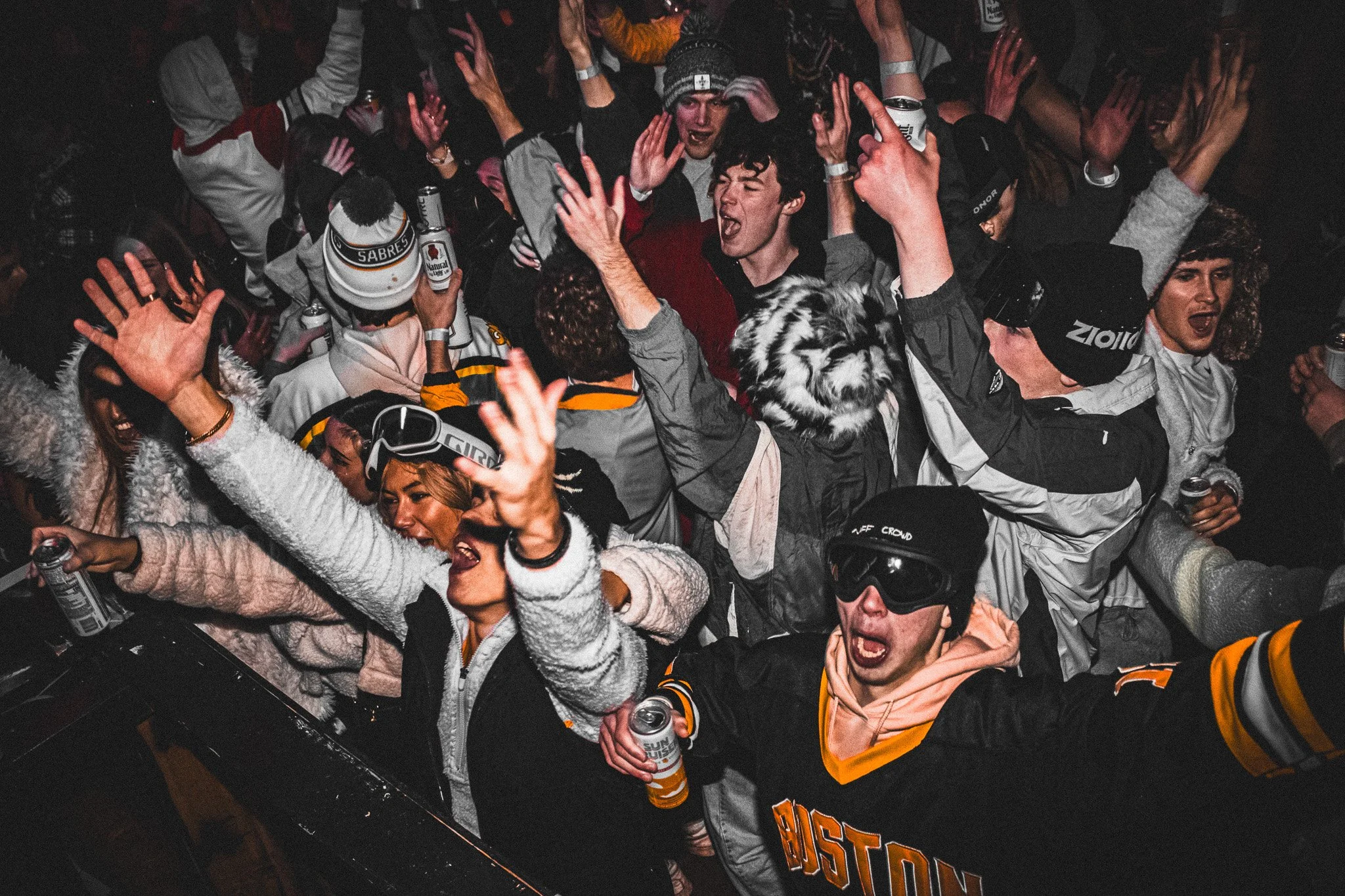 Group of young people at a lively party, dancing and celebrating with arms raised, some holding drinks, in a dimly lit indoor venue.