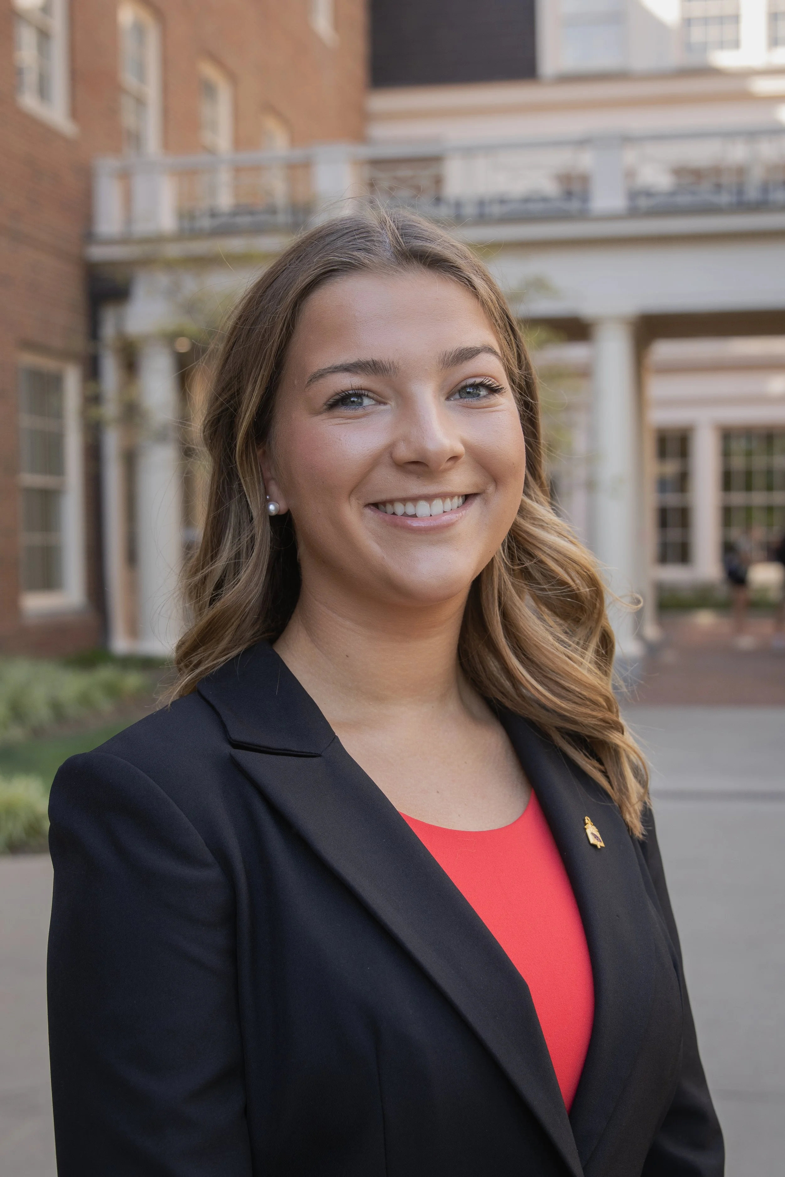 A young woman with long, wavy brown hair smiling outdoors in front of a brick and white building with columns, wearing a black blazer, red top, pearl earrings, and a small gold pin.