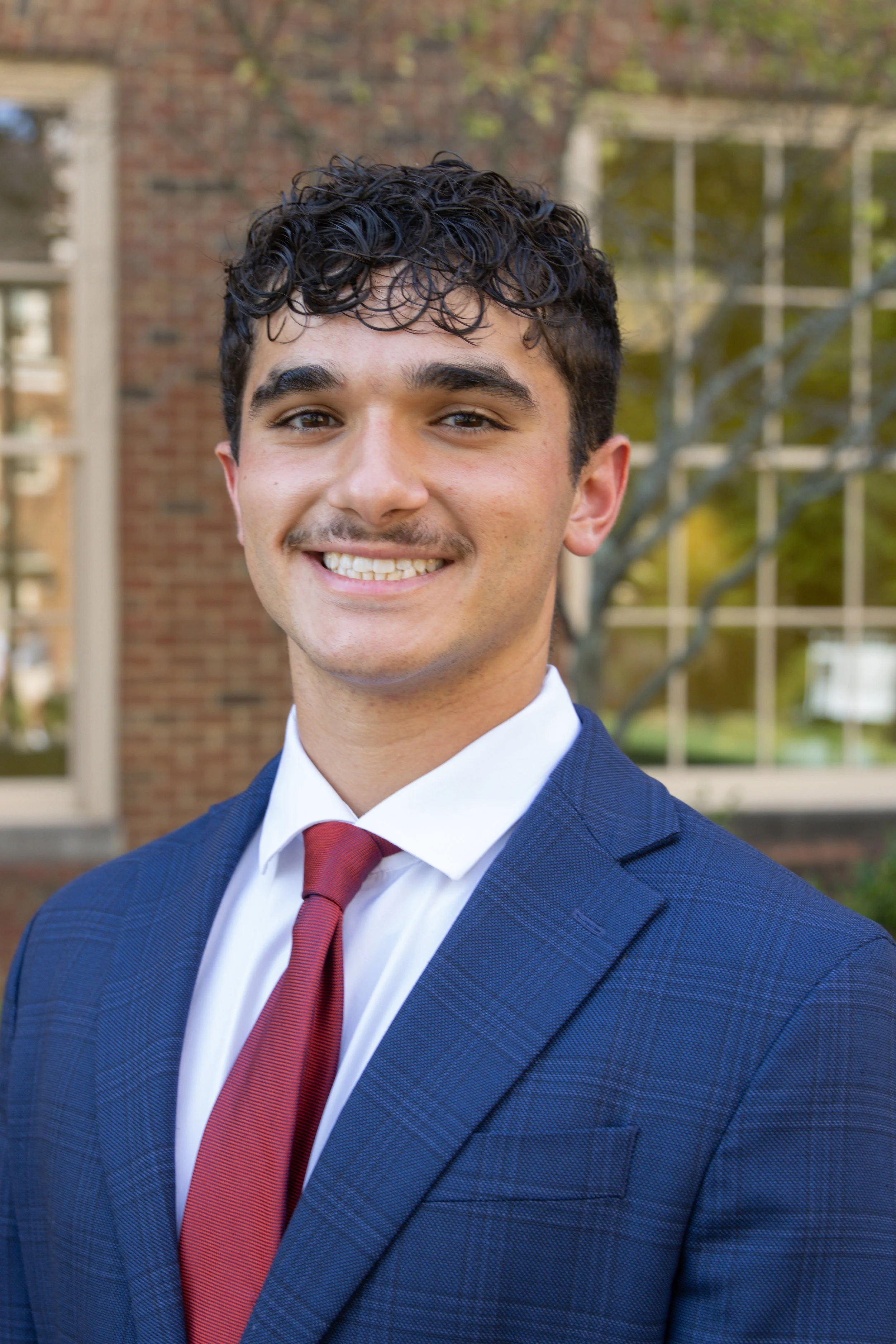 Young man outdoors in business attire smiling, with brick building and window panes in the background.