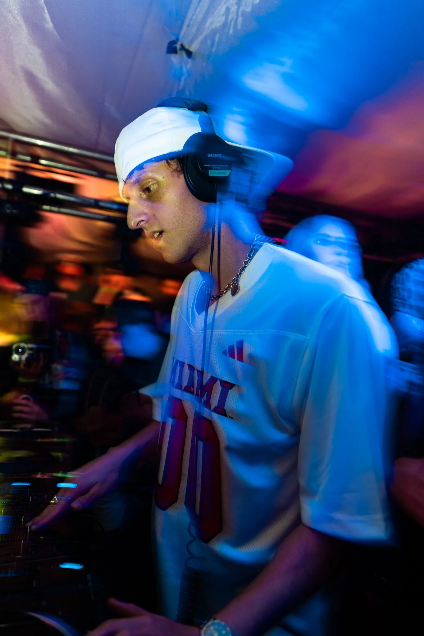 A DJ wearing a sports jersey, white cap, and headphones, performing at a crowded event with colorful lighting.