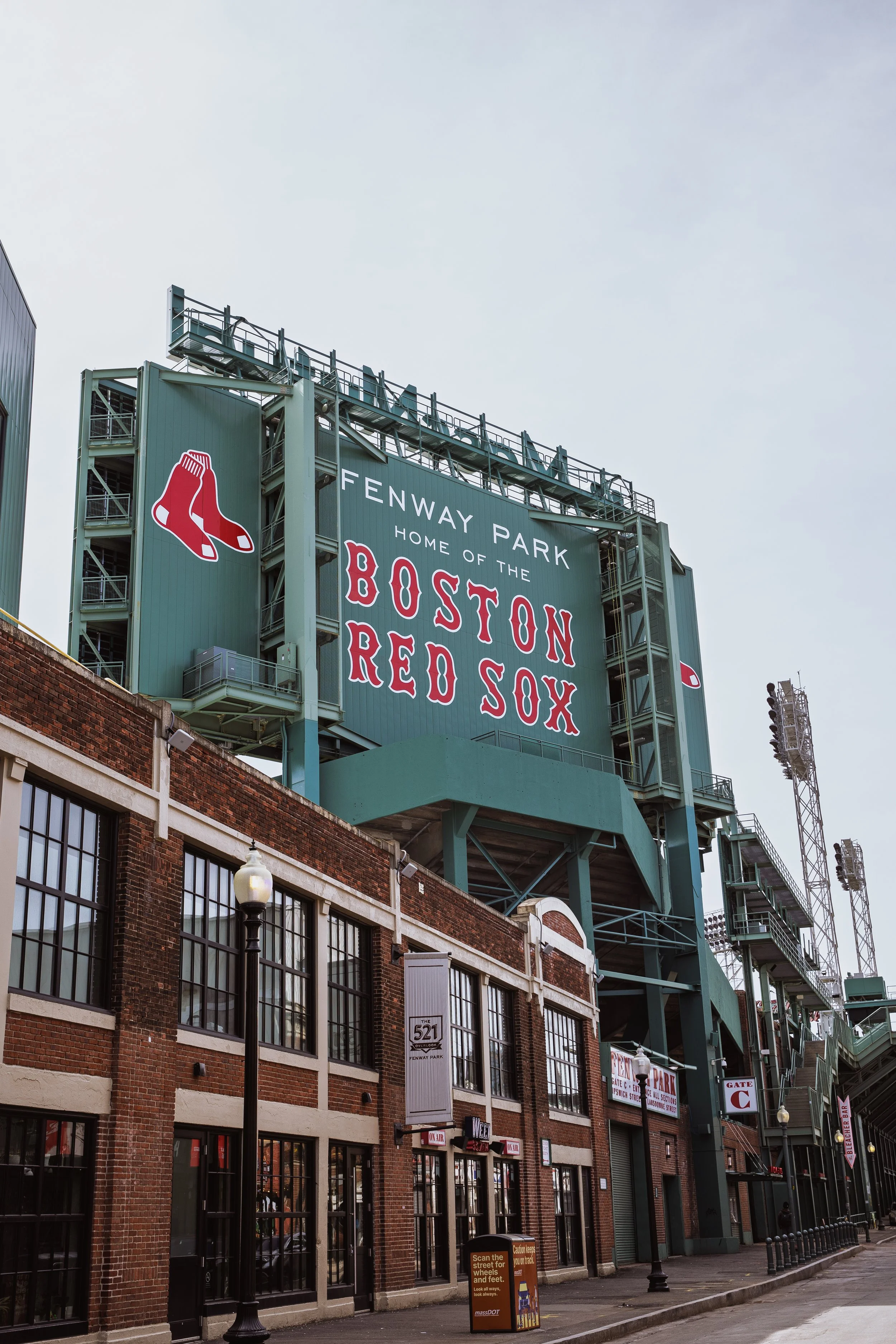 Exterior view of Fenway Park stadium in Boston, showing a large sign with the Boston Red Sox logo and text, and part of the brick building and sidewalk in front.