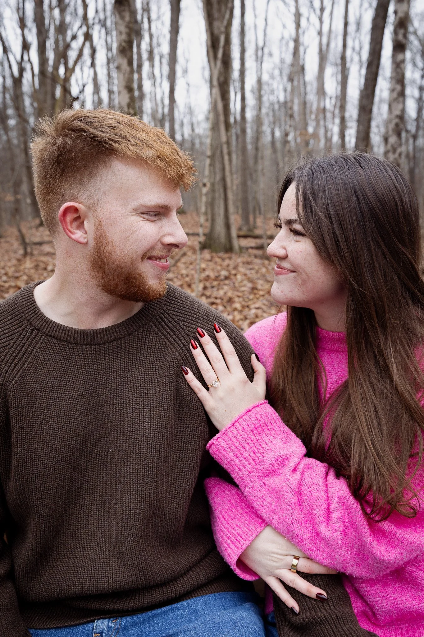 A happy couple looking at each other in a wooded area during fall, with leaves on the ground and trees in the background. The woman, wearing a pink sweater, has her hand on the man's chest, and they are smiling.