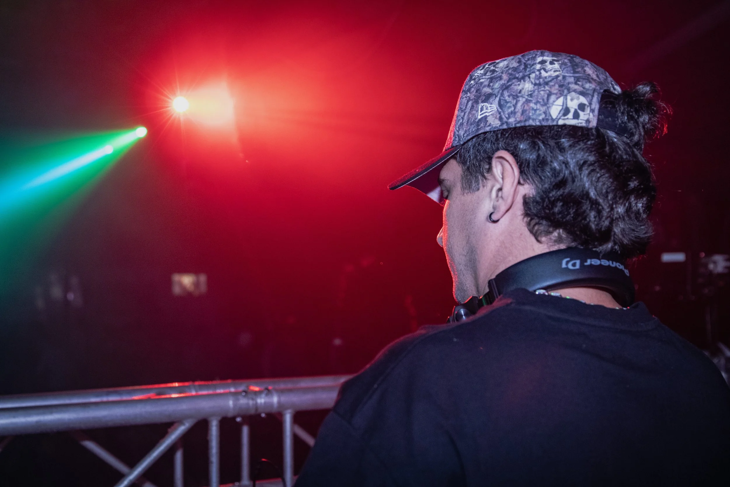 A person with long curly hair wearing a patterned baseball cap, earring, and headphones around neck, standing near a stage with colorful lights.