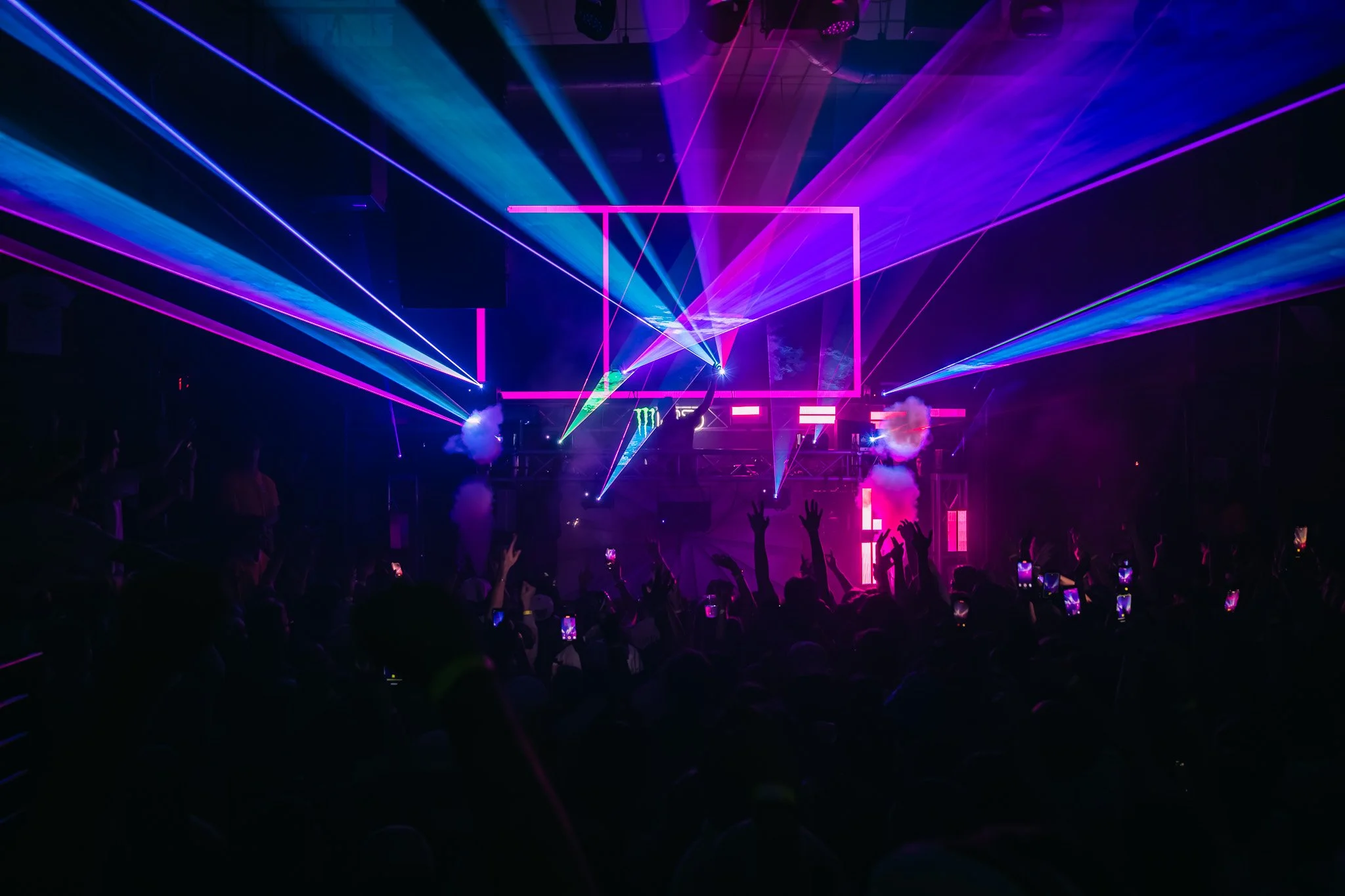 A concert scene with colorful laser lights and smoke effects, with a crowd of people raising their hands and capturing the moment on their devices.