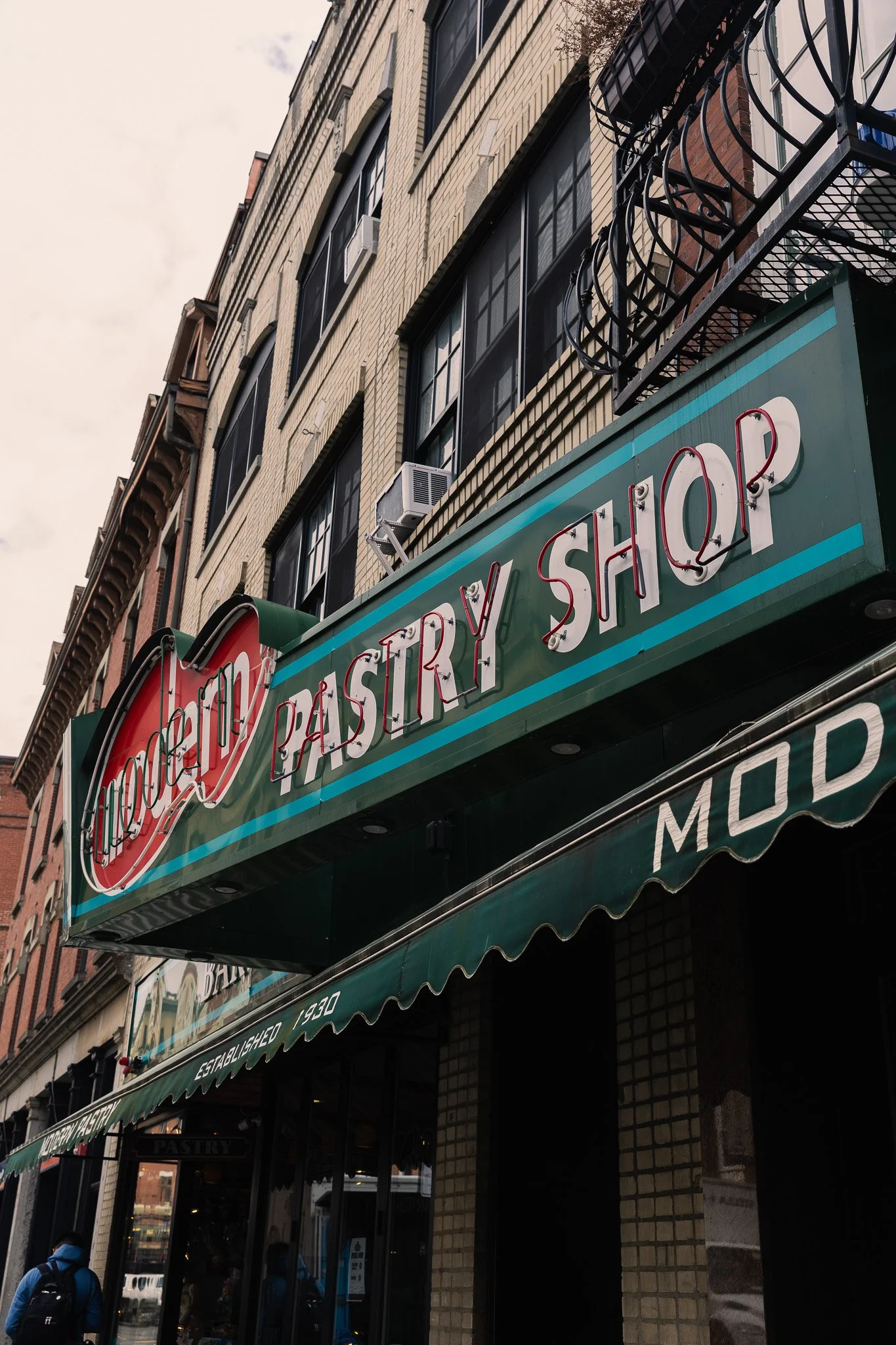 The exterior of a bakery and pastry shop with a large green awning, red and white signage, and people walking outside.