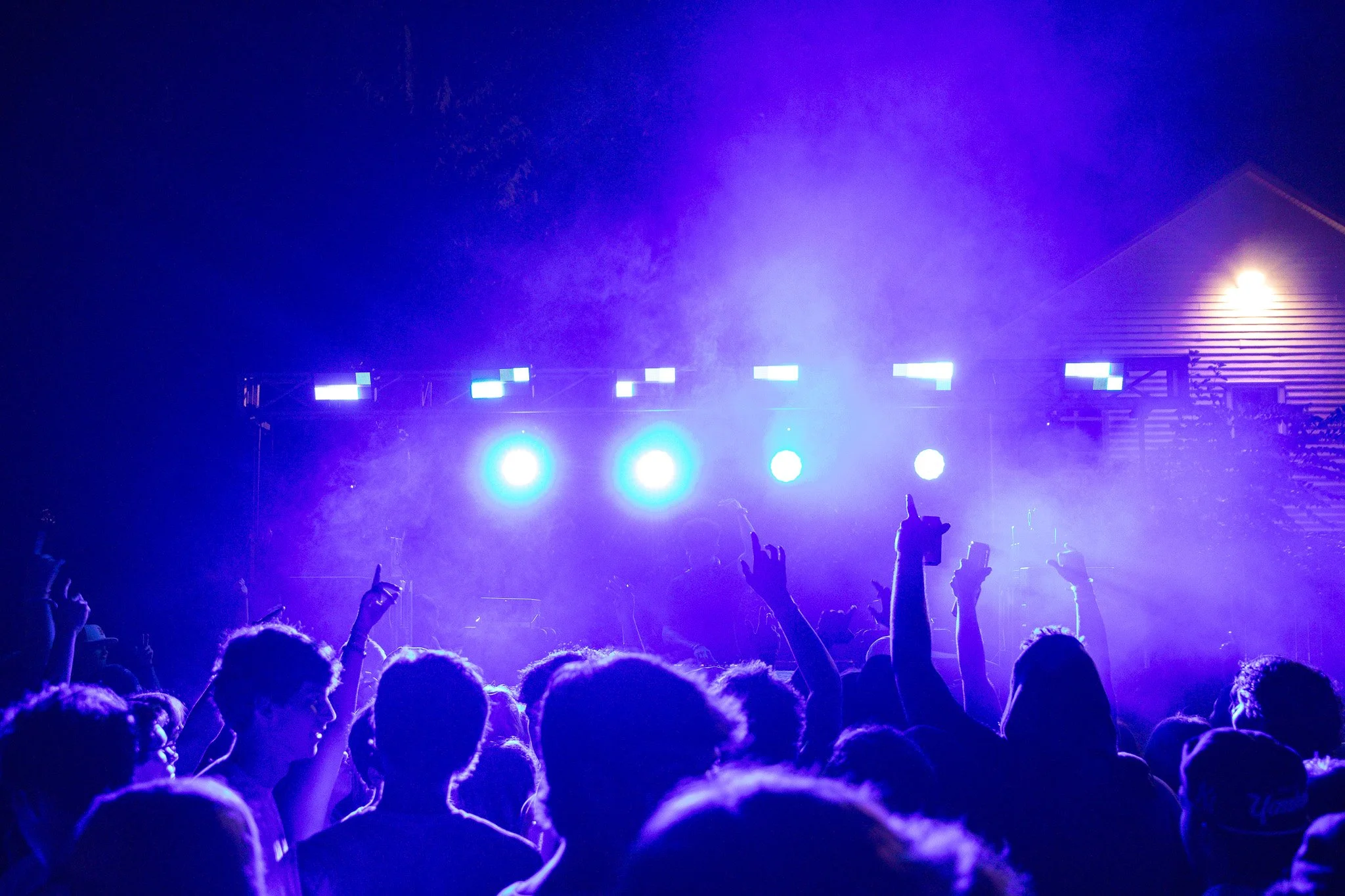 Crowd of people at a concert or music event with blue stage lights, some audience members raising their hands.